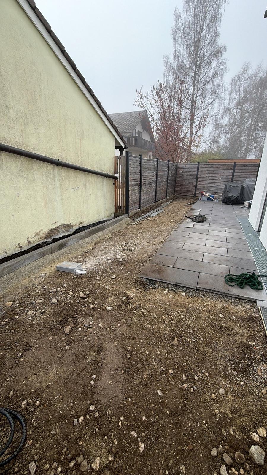 Gärtnerarbeiten im Garten, teilweise fertiggestellte Bodenplatten und unebener Boden, Zaun im Hintergrund, bewölkter Himmel.