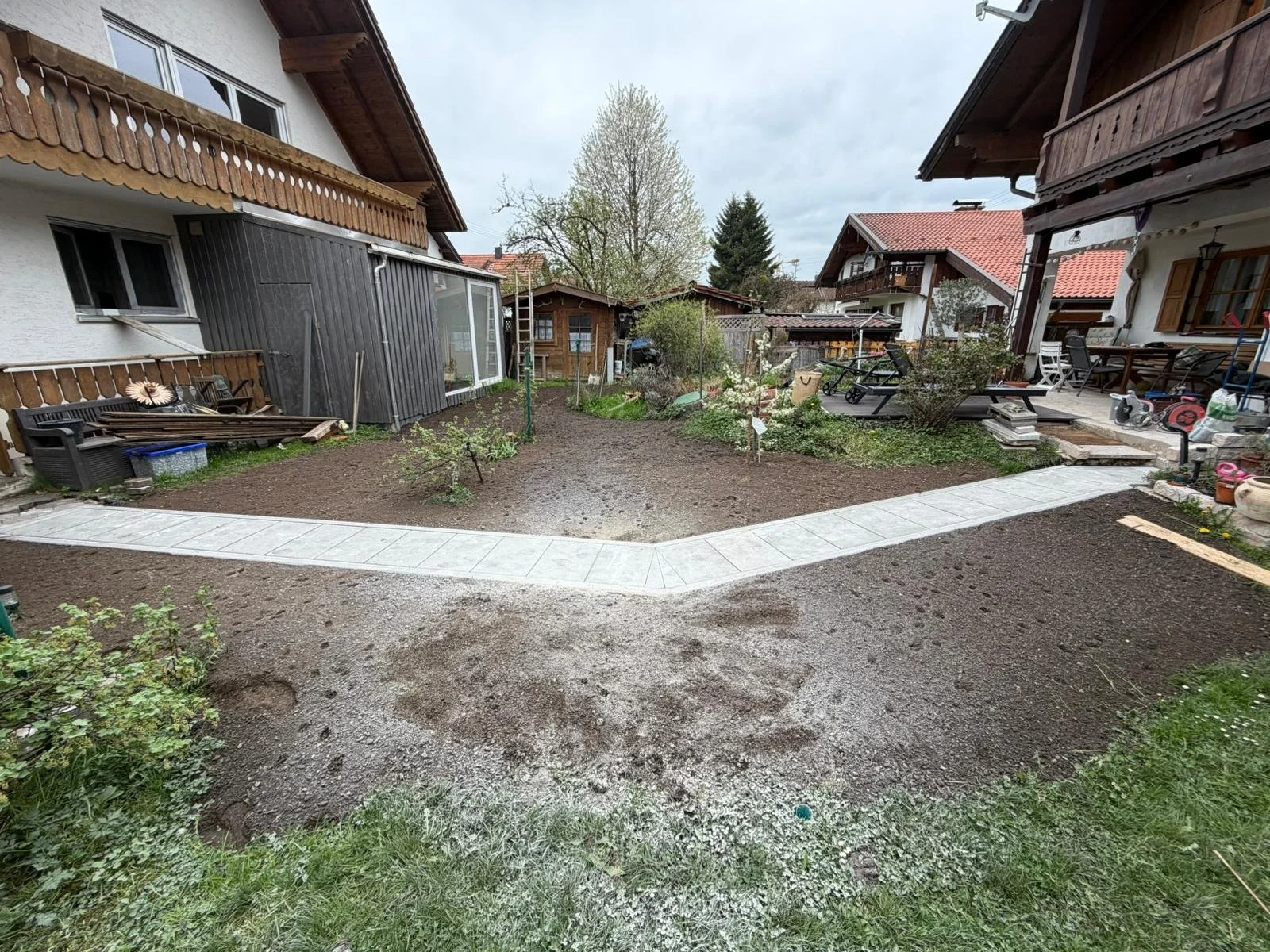 Garten mit verdichteter Oberfläche, im Hintergrund Bäume und Häuser mit Holzfassaden, im Vordergrund ein neu angelegter Weg aus Fliesen.
