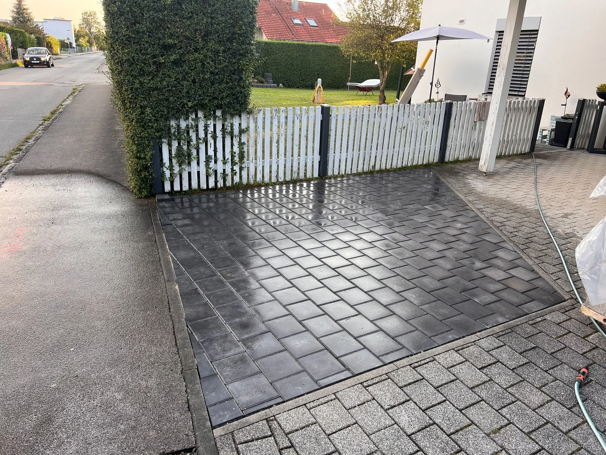 Garage mit schwarzen Pflastersteinen, umgeben von einer weißen Holzzaun und einem grünen Garten im Hintergrund.
