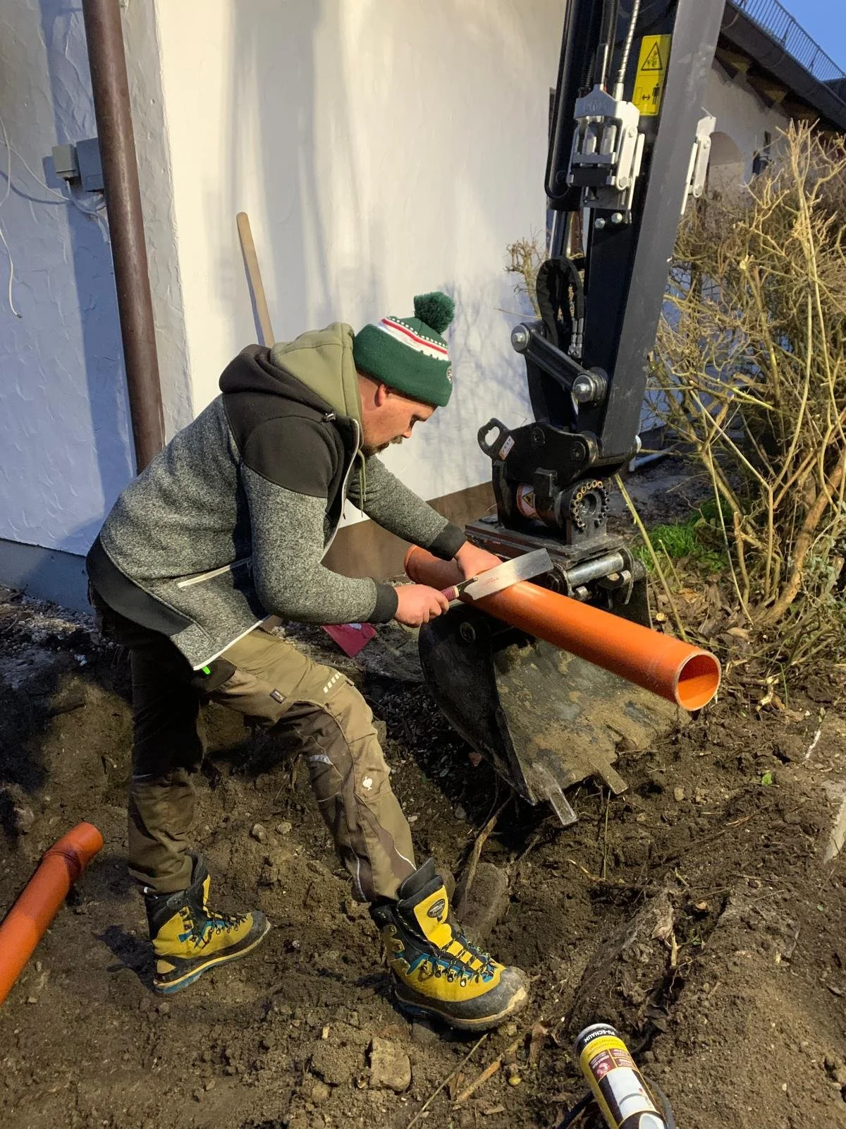 Mann arbeitet mit einer Bohrmaschine an einer Rohrinstallation im Boden vor einem Haus.