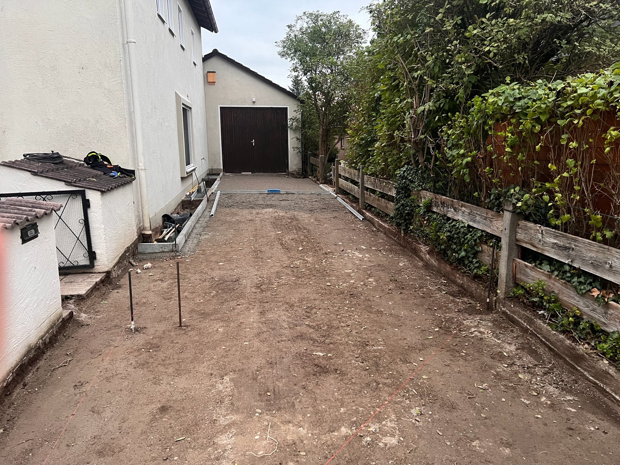 Gartenweg vor der Gartenarbeit, auf dem Boden liegt Erde, im Hintergrund ein Haus mit Garage, rechts ein Zaun mit Büschen, links Hauswand mit Fenster.