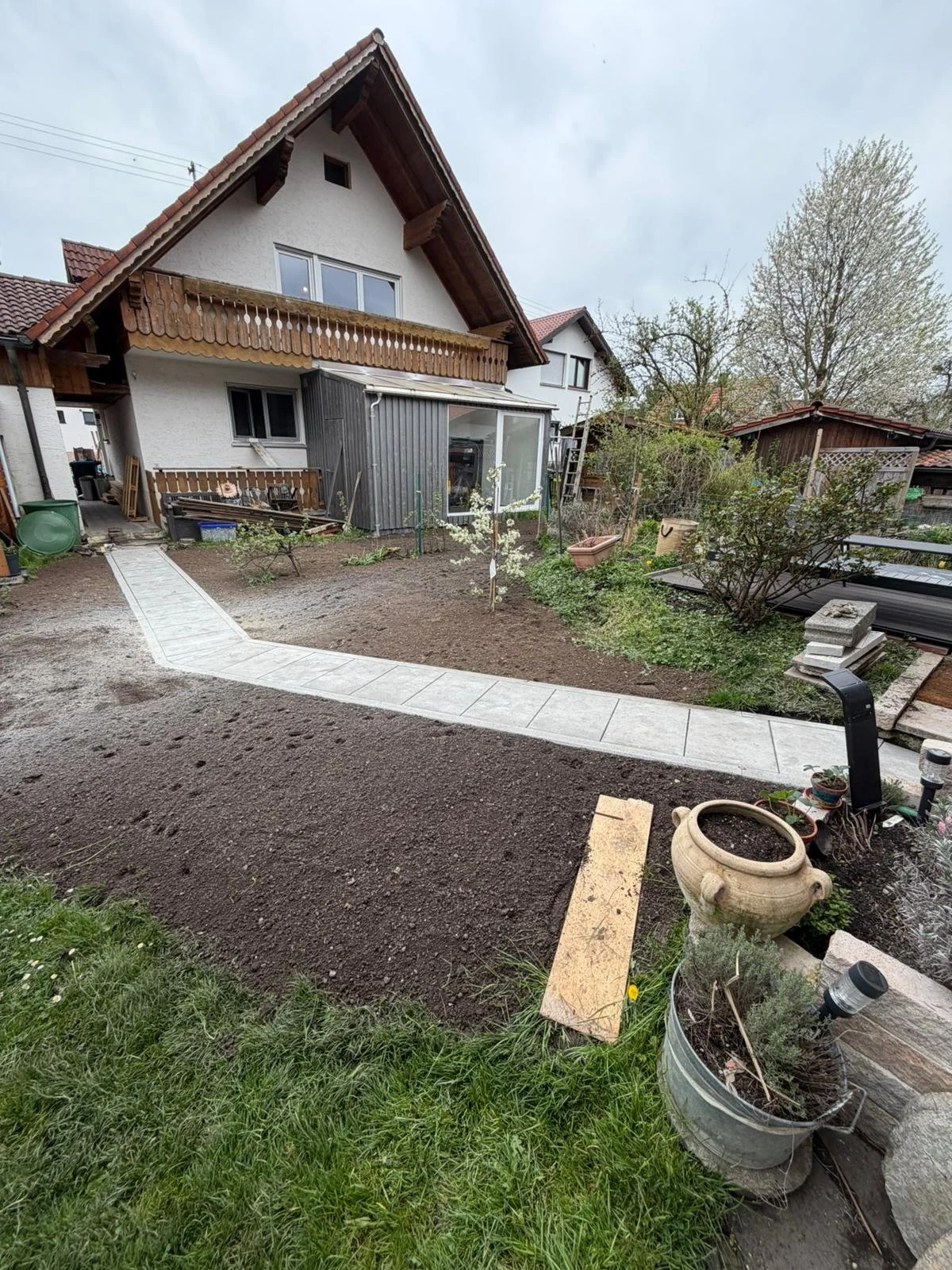 Gärtnerarbeiten im Vorgarten eines Hauses, mit frisch gepflastertem Weg, bepflanztem Bereich und Gartenmöbeln, im Hintergrund das Haus mit Balkon