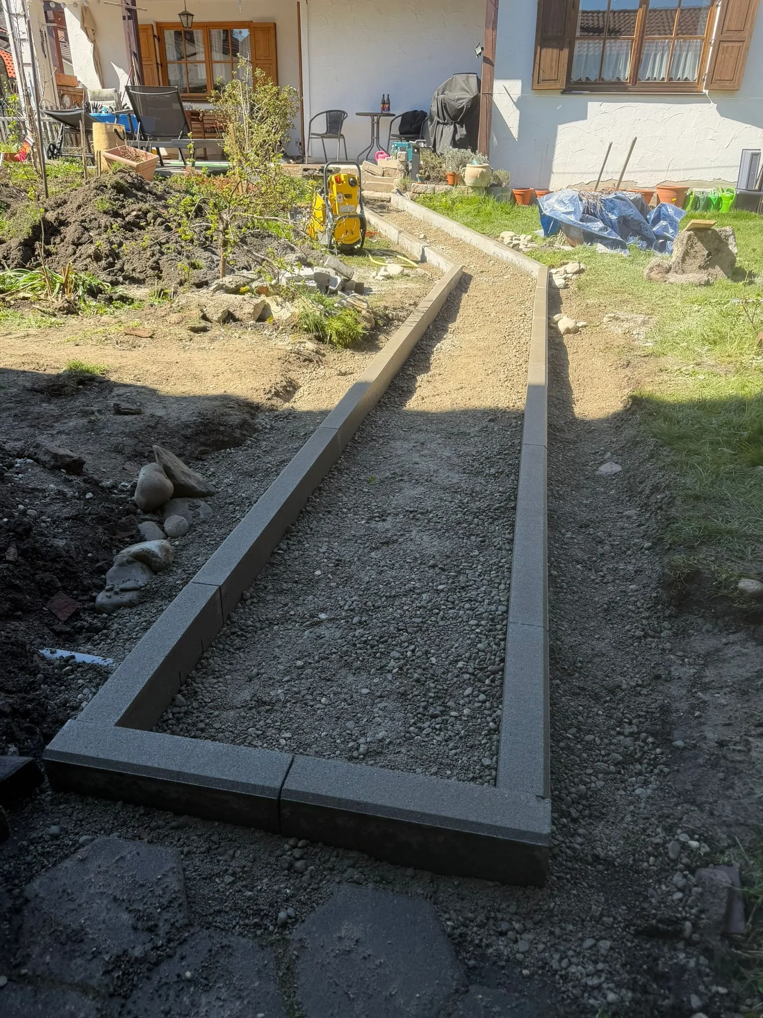 Der Garten wird neu gestaltet, mit einem frisch gepflasterten Weg und einer Blumenrabatte im Vordergrund, sowie Gartenmöbeln und Pflanzen im Hintergrund.