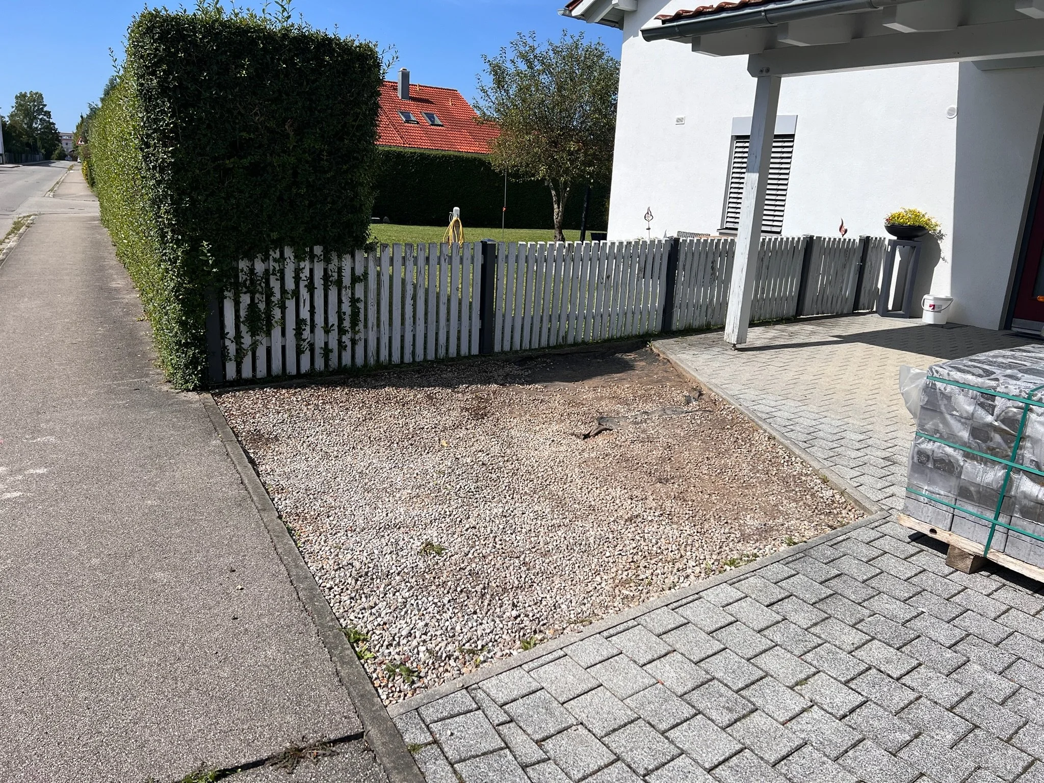 Ein Vorhof mit gepflastertem Gehweg, reserviertem Parkplatz mit Kies und einem weißen Haus mit Terrasse, umgeben von einem Zaun und einem grünen Garten im Hintergrund.