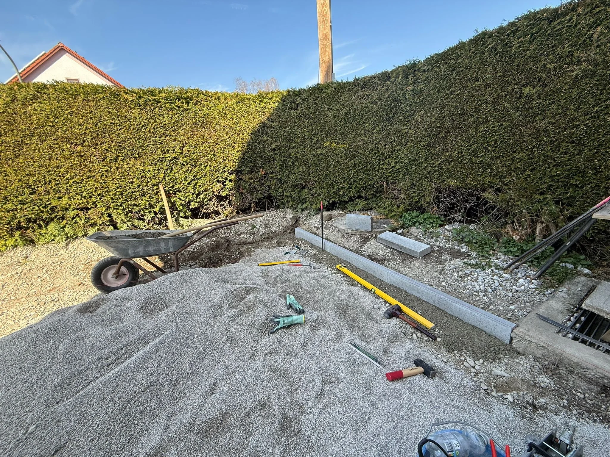 Baustelle mit Sand, Werkzeug und Baustoffe, vor einer grünen Hecke und einem Wohnhaus im Hintergrund.