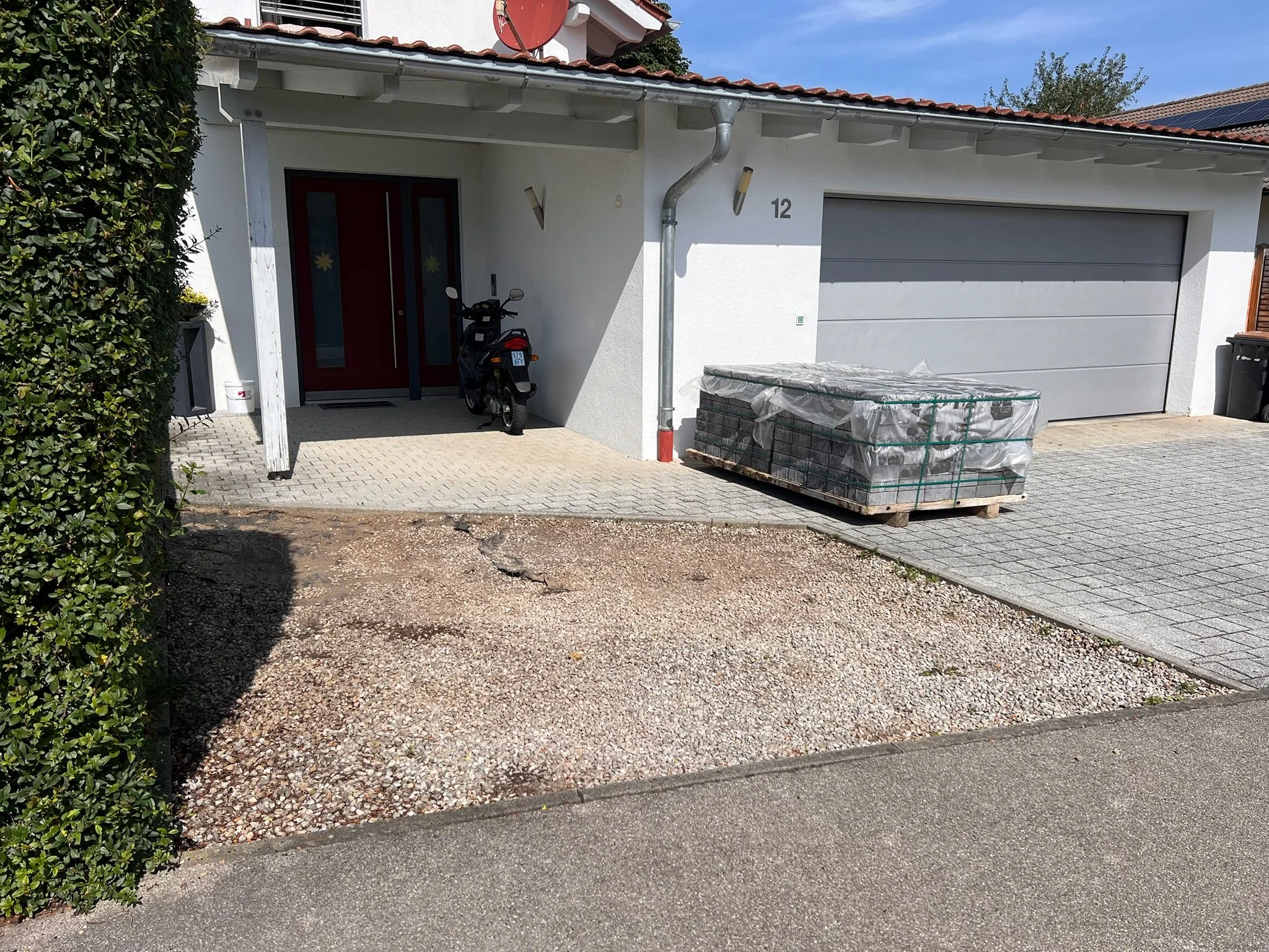 Ein Haus mit weißer Fassade, grauer Garagentür und rotem Doppelfenster. Neben der Tür steht ein schwarz-rot-weißes Moped. Auf einer Palette vor der Garage sind Steine oder Ziegel, die verpackt sind. Der Eingangsbereich ist teilweise überdacht. Es gib