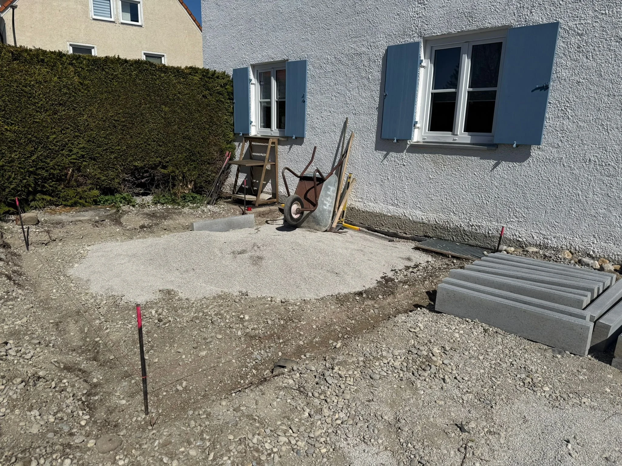 Baustelle vor einem Haus mit zwei offenen Fenstern und angelegtem Fundament für eine Terrasse oder einen Eingangsbereich, mit Steinen und Baumaterialien vorbereitet.