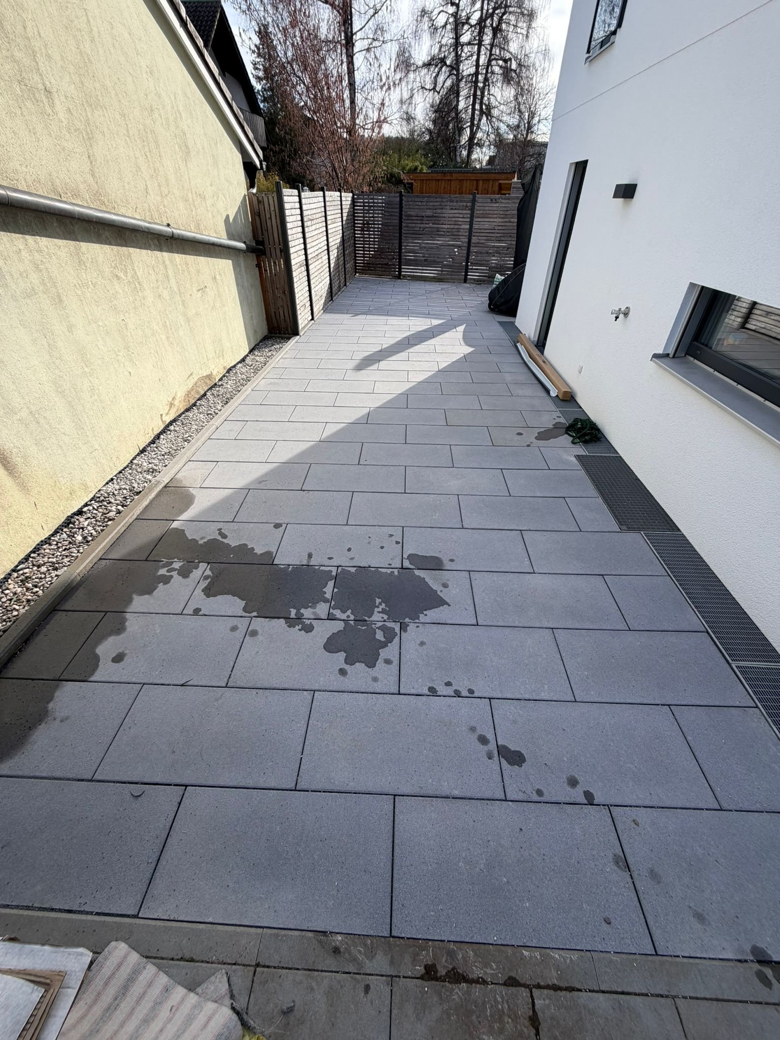 Neuer Terrasse mit großen grauen Fliesen, Wasserflecken und einem winterlichen Garten im Hintergrund, Blick von unten auf die Terrasse.