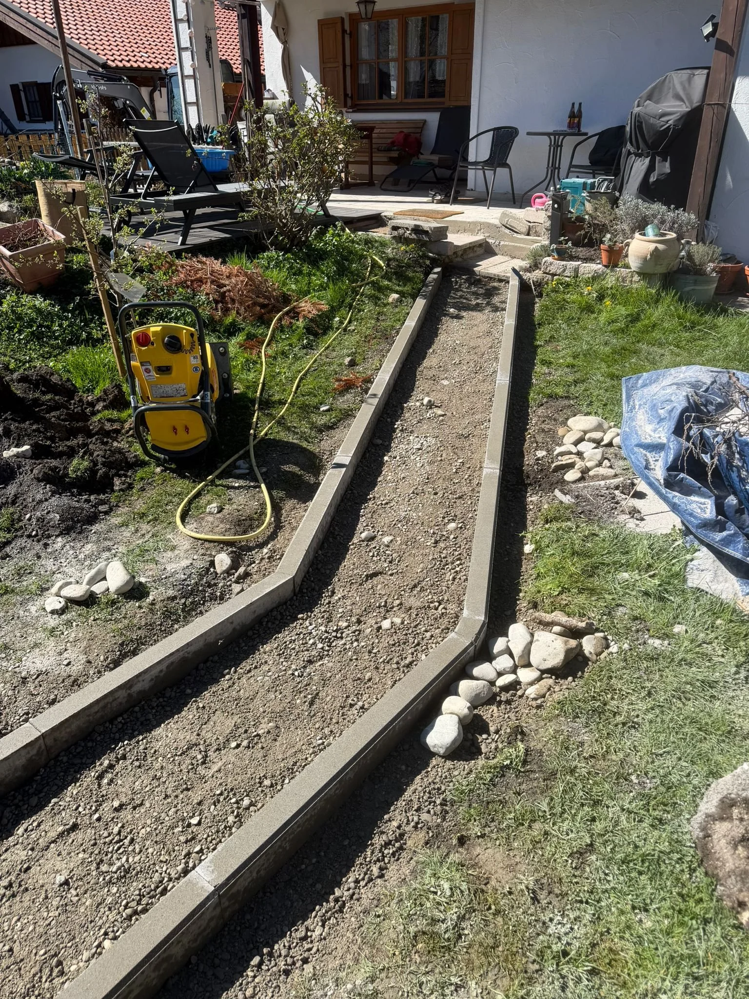Gärtner arbeitet an einem Wegefundament aus Beton im Garten, umgeben von Pflanzen, Möbeln und Gartengeräten.