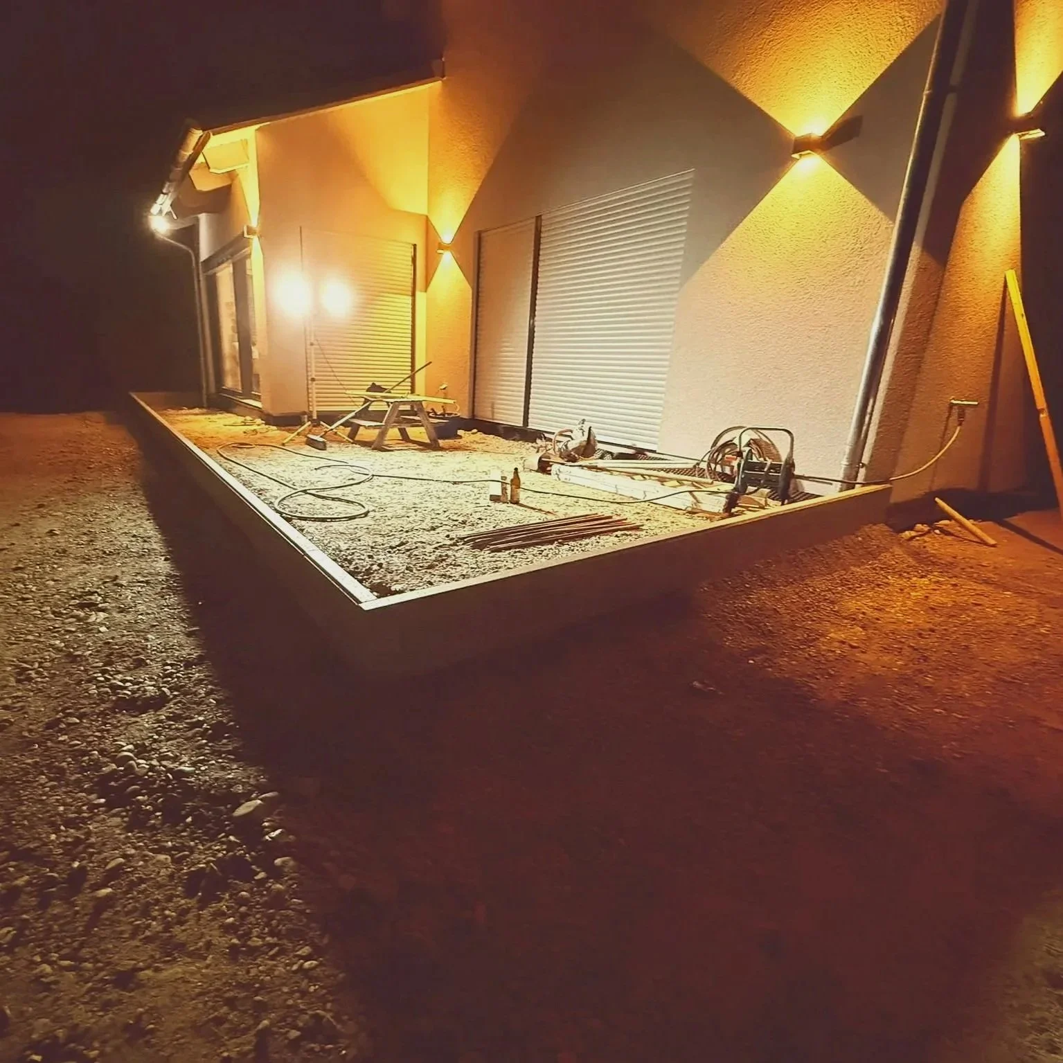 Baustelle bei Nacht mit eingeputztem Terrassenboden vor einem Haus, das Fenster mit Rollläden hat, und Baugeräten sowie Beleuchtung