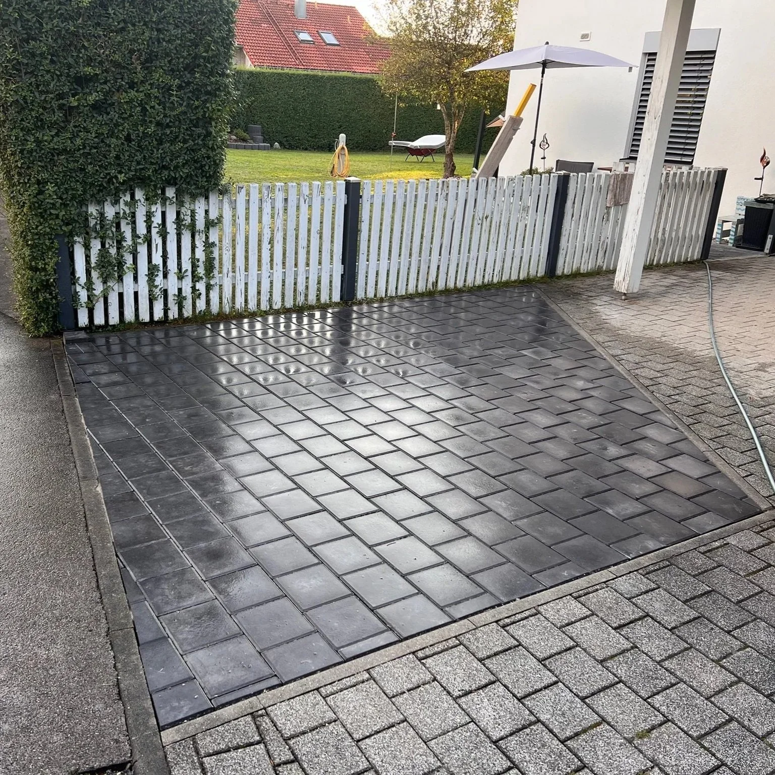 Ein neu gepflasterter Autostellplatz mit schwarzen Steinen, umgeben von einem gepflasterten Bereich. Im Hintergrund ein weißer Lattenzaun, grüne Bäume und ein Garten mit Rasenflächen und Gartenmöbeln.