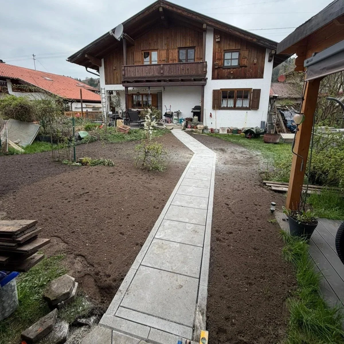 Garten mit Gehweg, unbedeckter Boden, Haus mit Holz- und weißen Wänden, Gartenterrasse, Gartenmöbel, Pflanzen, Gartengeräte und Werkzeuge.