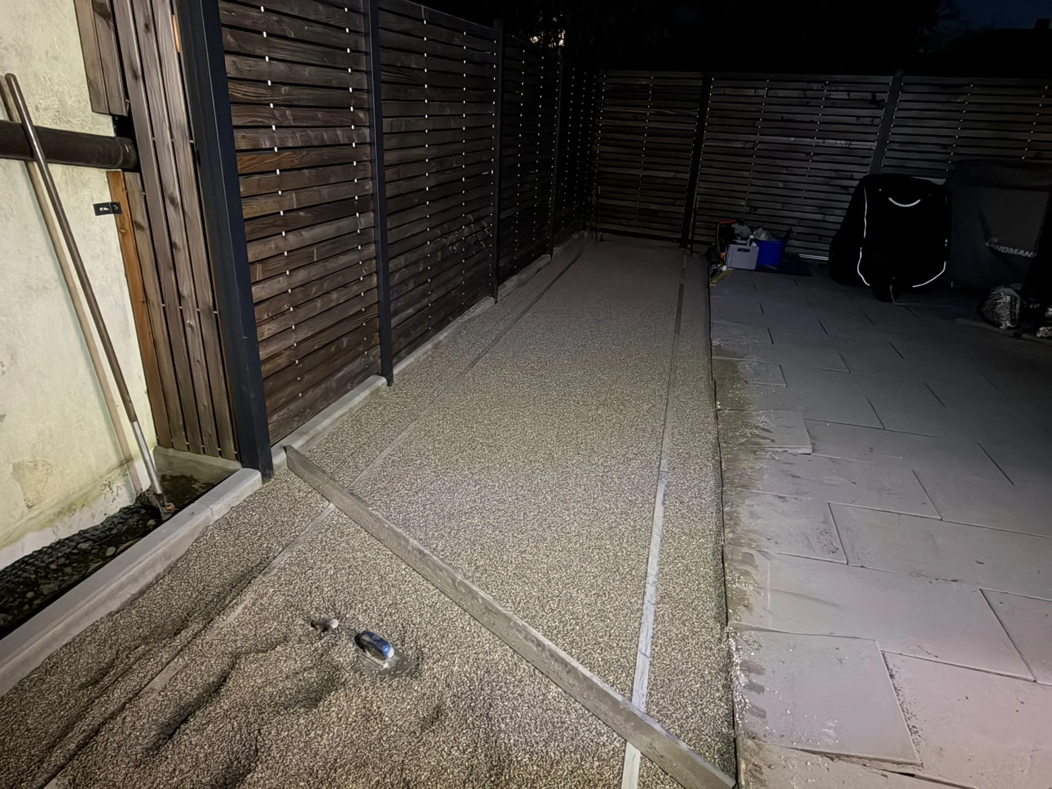 Außenbereich im Dunkeln mit Holzlattenzaun, Arbeitsmaterialien und Equipment auf dem Boden, teilweise ausgelegt.