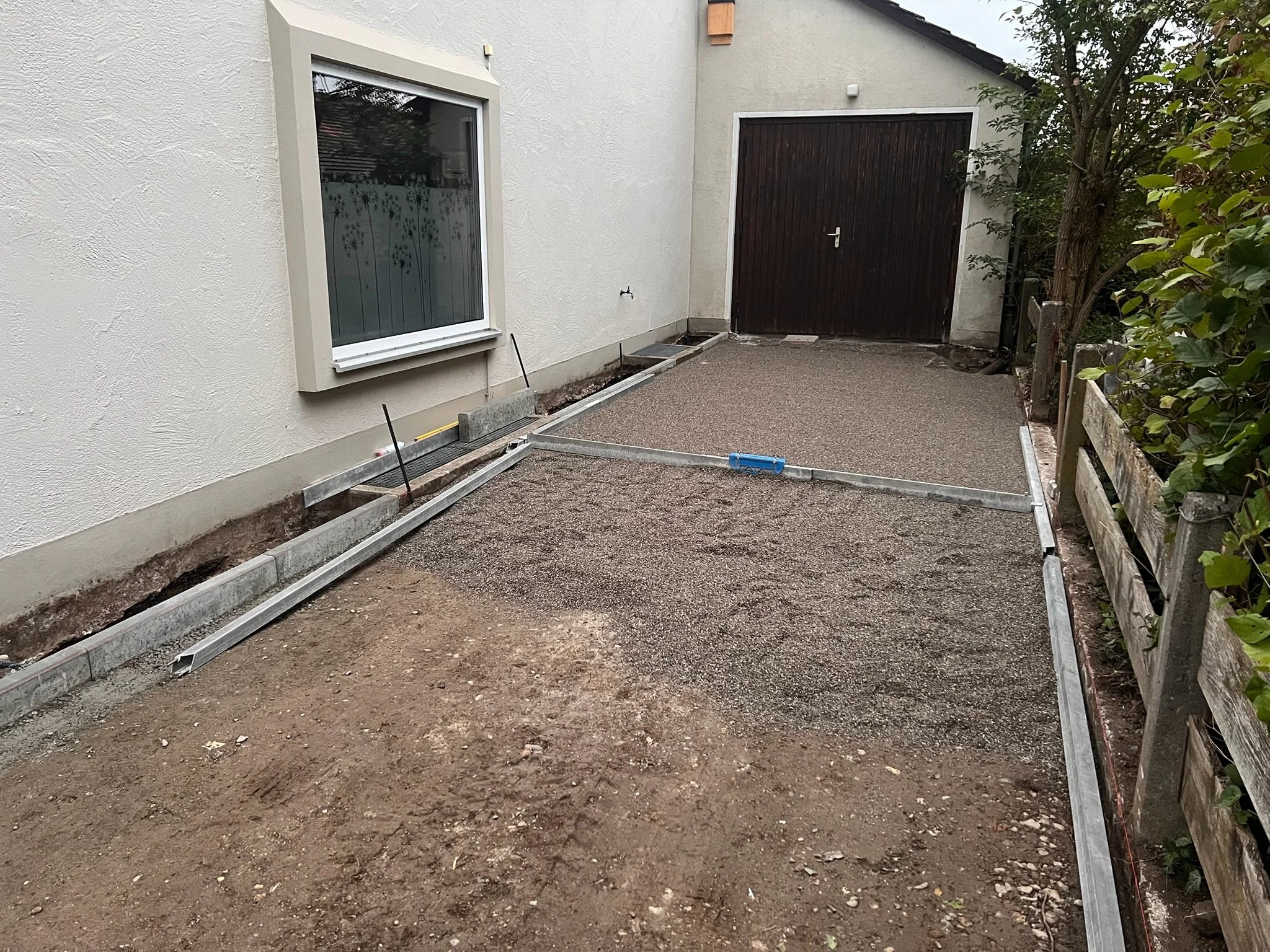 Der eingereichte Bild zeigt eine Zufahrt, die mit Pflastersteinen vorbereitet wird, mit einem weißen Haus an der linken Seite und einem lackierten Holztor am Ende der Zufahrt.