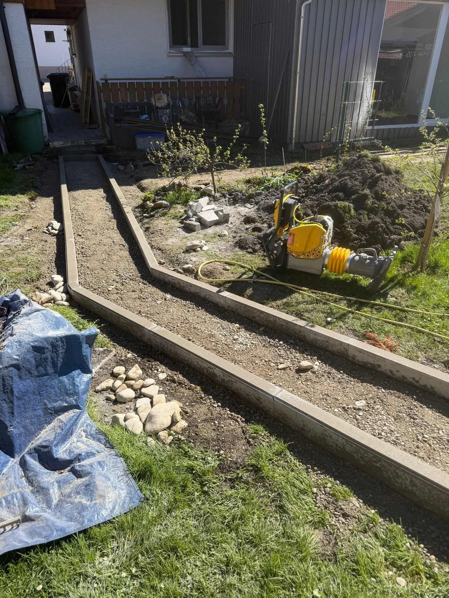 Ein Gartenbereich mit einem neuen Pflasterweg, einem kleinen Baum, Gartenwerkzeug und Baustoffen auf der Wiese. Im Hintergrund ist ein Haus mit Fenster und einer Holzveranda.