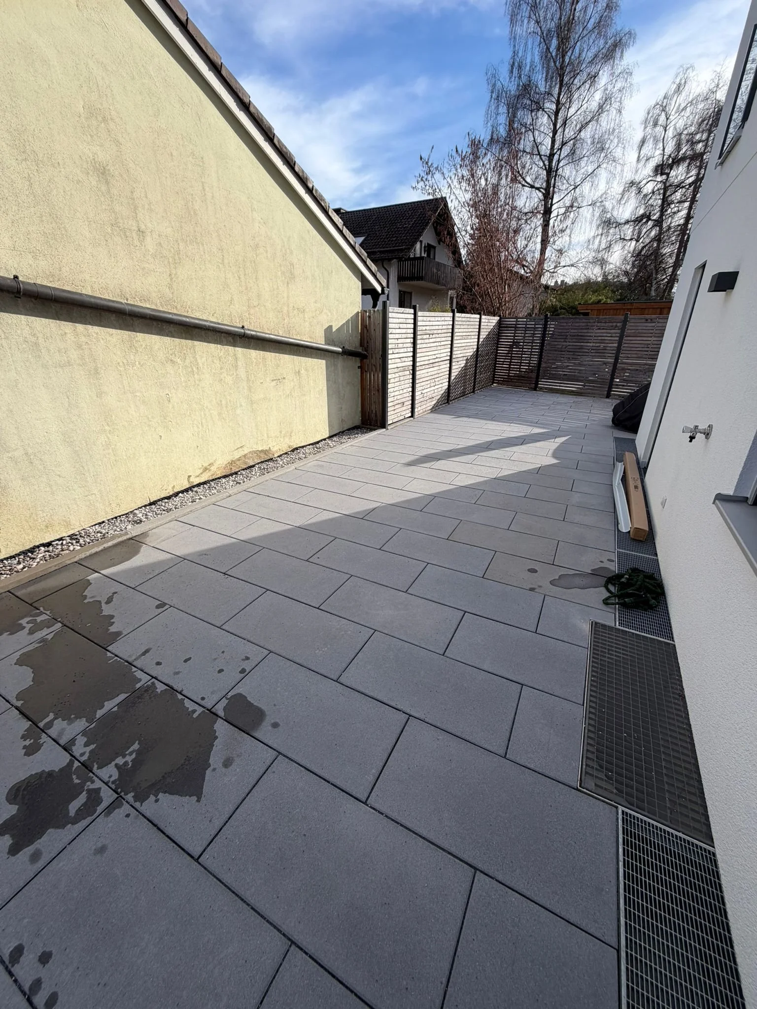 Ein geplanter Bereich für Pflasterarbeiten mit gepflasterten Steinen, umgeben von Kies und Gartenpflanzen, vor einem weißen Haus mit blauen Fensterläden.
