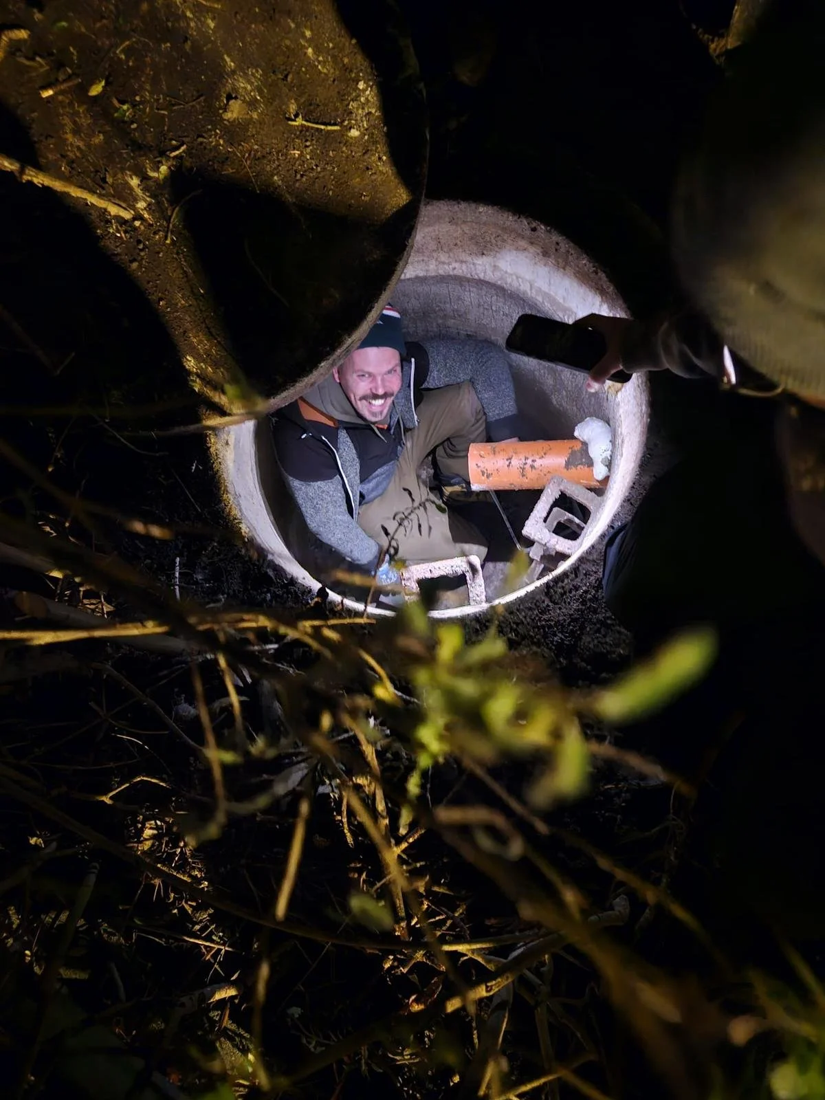 Ein Mann sitzt in einer künstlerischen menschlichen Trinkwasserleitung, umgeben von Pflanzen, im Dunkeln, mit einem Lächeln auf dem Gesicht.