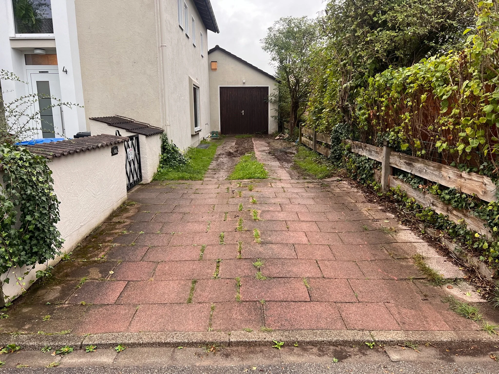 Ein Pflasterweg führt zu einer weißen Hausfassade mit Garage im Hintergrund. Links und rechts sind Gärten mit Büschen und Bäumen, einige Pflanzen wachsen zwischen den Pflastersteinen.