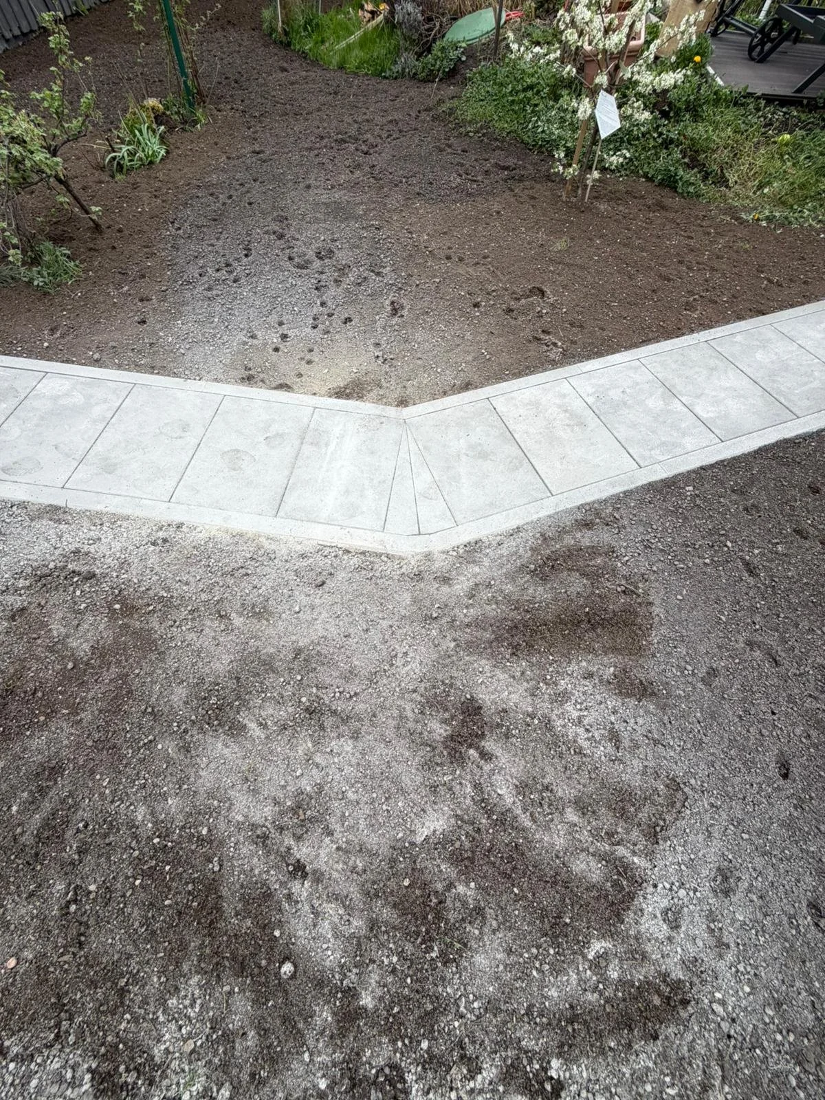 Gepflasterter Weg an der Grenze zu aufgeworfener Erde, umgeben von Gartenflächen mit Pflanzen und Blumen.
