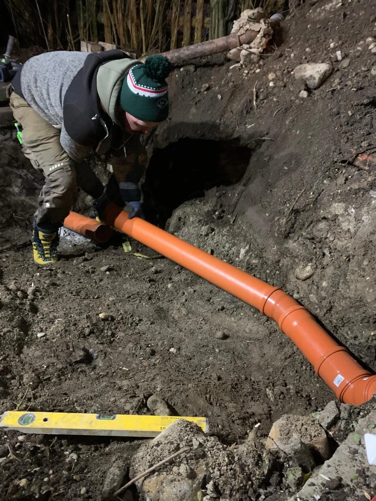 Eine Person installiert eine orange Rohrleitung in einem erdigen Graben, vermutlich für die Abwasser- oder Wasserleitung.