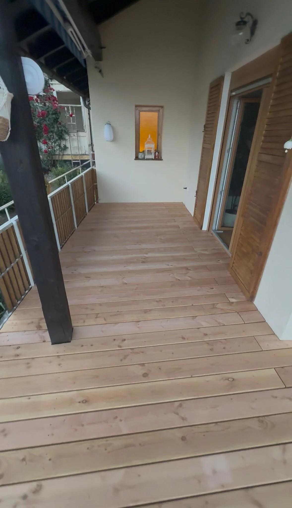 Ein Balkon mit Holzboden und einer Markise, mit einer Tür und einem Fenster, draußen sind Blumen und eine Pergola sichtbar.