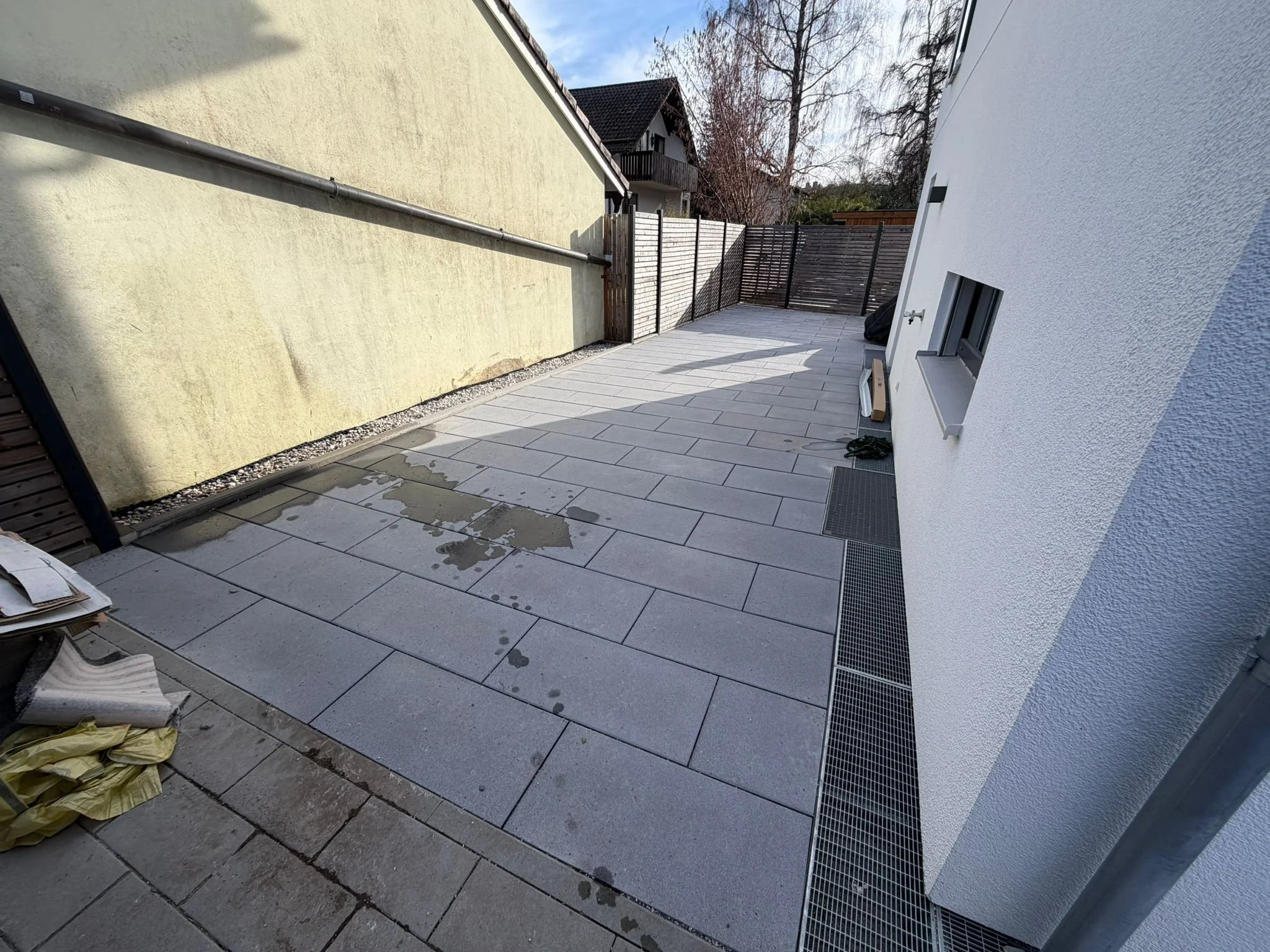 Ein angrenzender Außenbereich eines Hauses mit einer Terrasse aus großen, hellgrauen Fliesen, schwarzen Gitterabläufen am Rand und einer weißen und einer gelben Wand. Es liegen einige Gegenstände und Papiere auf der Terrasse, im Hintergrund sind Bäum