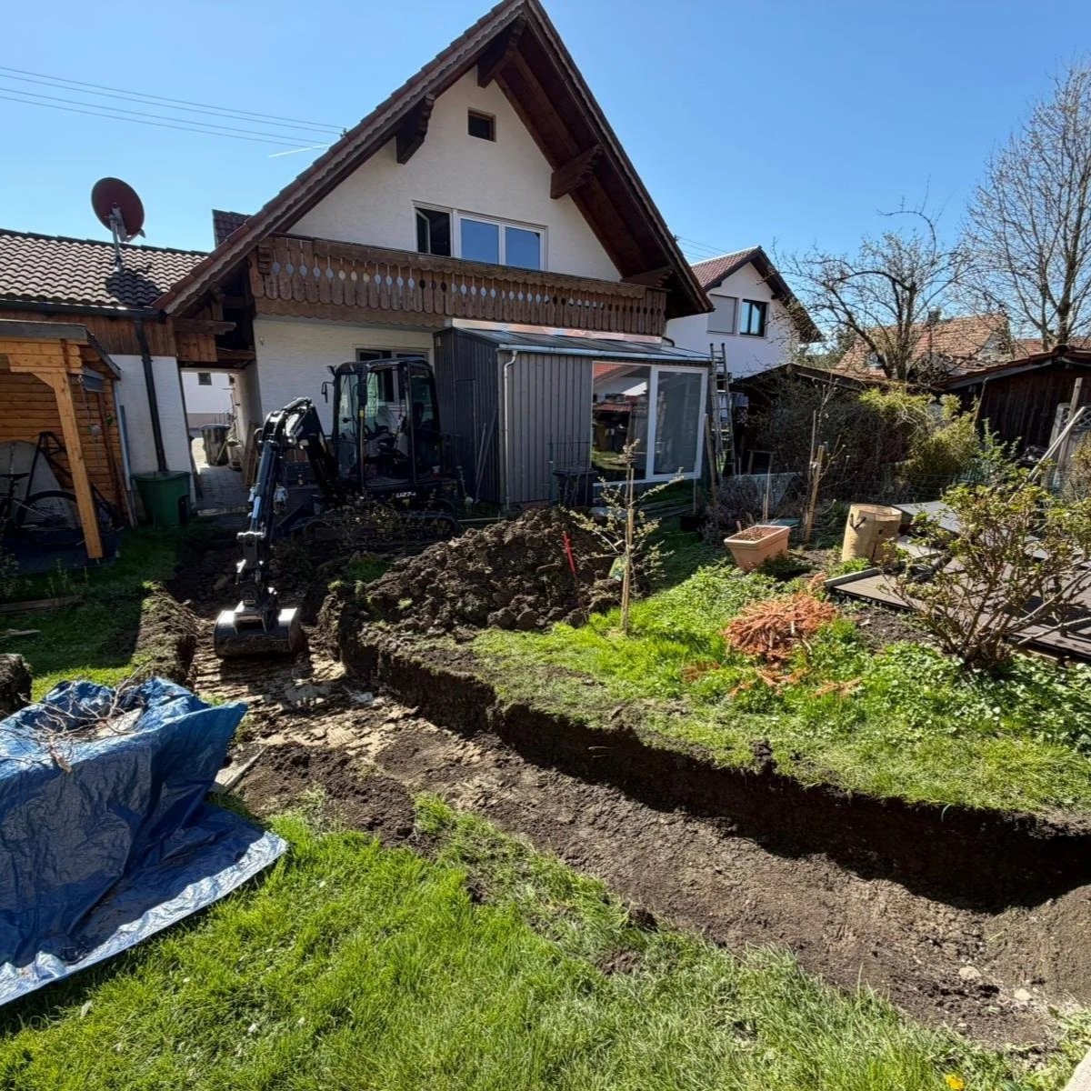 Gartenarbeiten im Hinterhof eines Hauses, Baustelle mit Erde und einem kleinen Bagger, im Hintergrund ein Haus mit Balkon und Garten, sonniger Tag.
