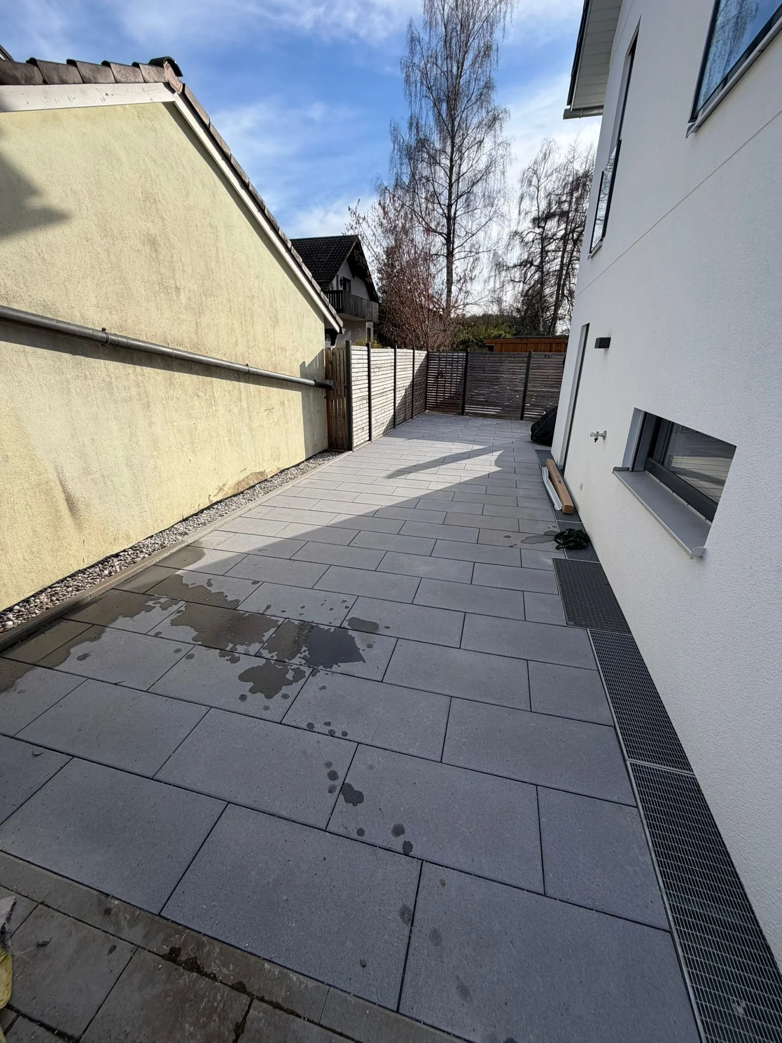 Ein moderner Innenhof mit großen Pflastersteinen, Wasserflecken und einem weiß gestrichenen Haus mit mehreren Fenstern. Es gibt einen Holz- oder Metallzaun im Hintergrund und eine klare, blaue Himmel mit vereinzelten Wolken.