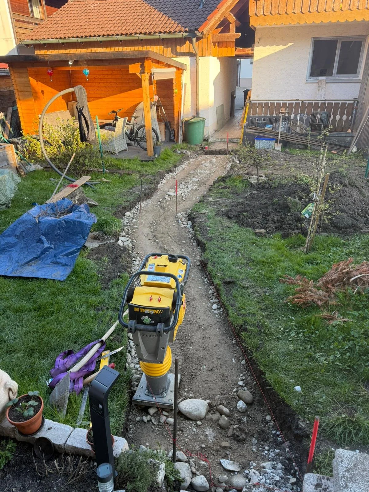 Ein Garten im Bau mit einer Gehwegfundamentmarkierung, betoniertem Weg und Gartenpflanzen. Es gibt Werkzeuge, einen Rasenmäher und eine Baustation mit Kabeln.
