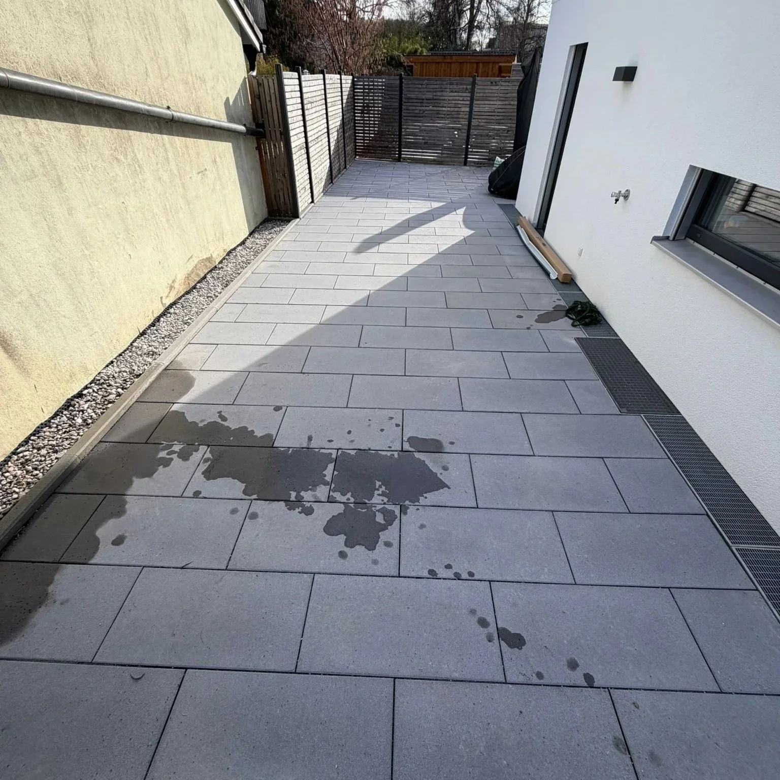 Ein moderner Balkon mit grau gefliesten Bodenplatten, einem weißen Wand, einem dunklen Holzzaun im Hintergrund, einigen Gegenständen am Rand und Wasserflecken auf den Fliesen.