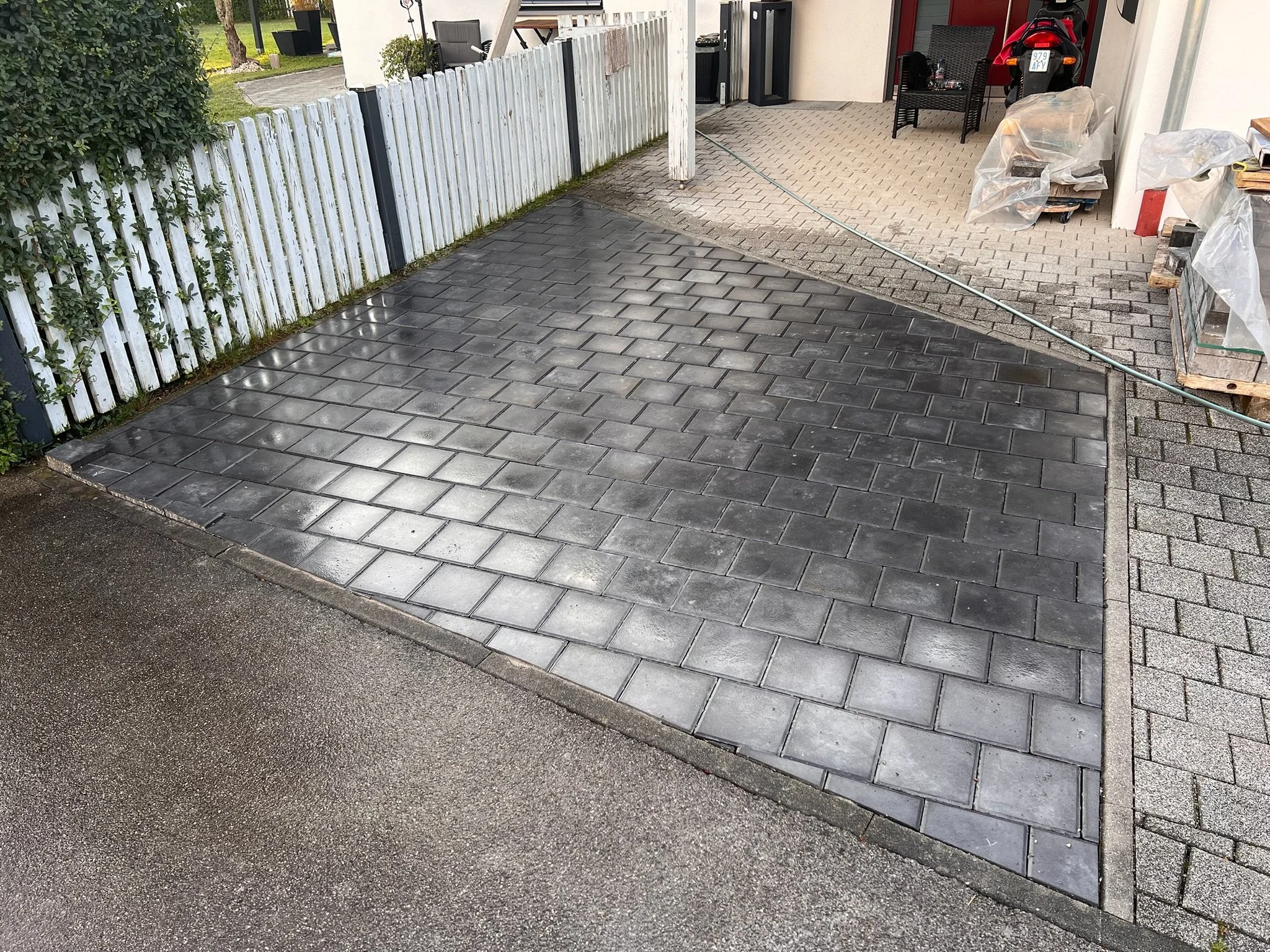 Neue schwarze Pflastersteine im Vorgarten, vor einem weißen Holzzaun, neben einer Einfahrt, im Hintergrund ein Sitzbereich mit Stühlen und eine Garage mit Werkzeugen und Materialien.
