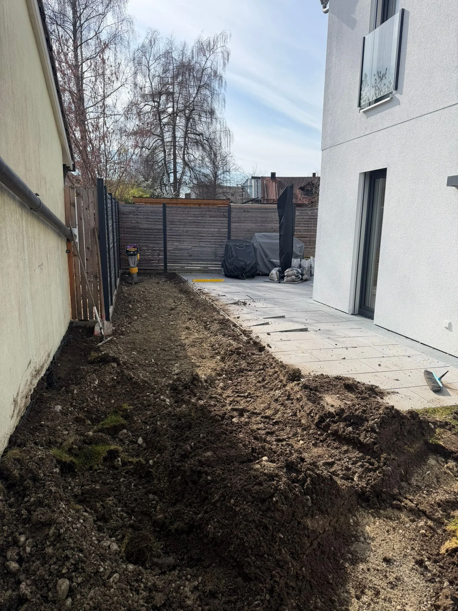 Ein Gartenbereich mit ungepflastertem Boden auf der linken Seite und einer gepflasterten Terrasse auf der rechten Seite, im Hintergrund eine Holzzaun und teilweise Bäume ohne Blätter.