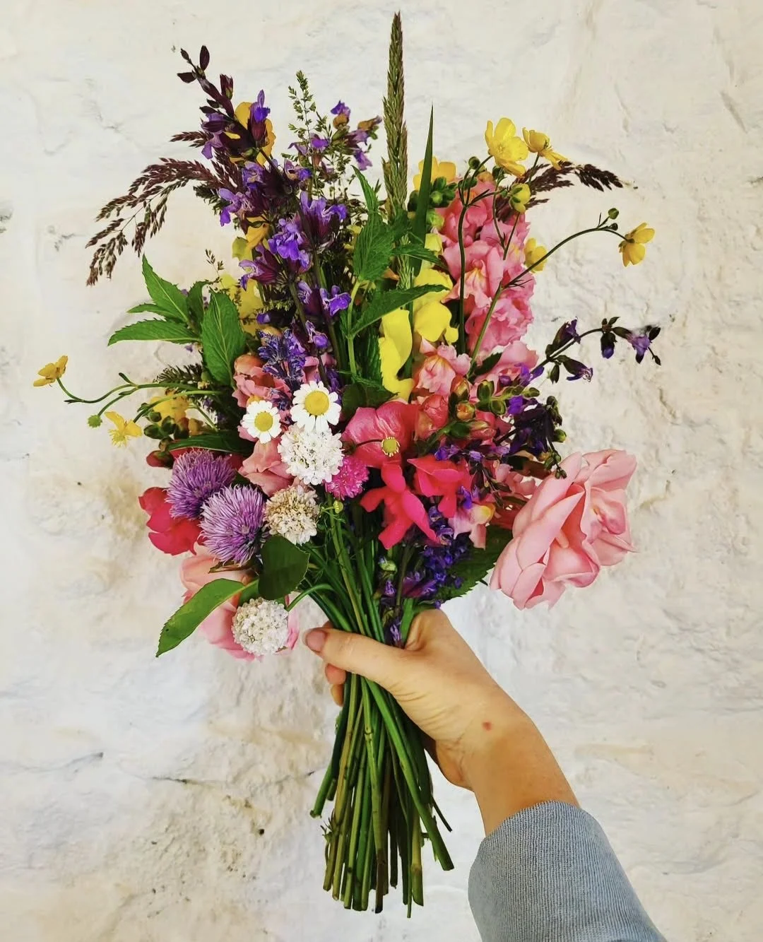A colorful bouquet of mixed flowers including pink roses, yellow and purple blooms, white daisies, and various green foliage being held up against a plain white wall.
