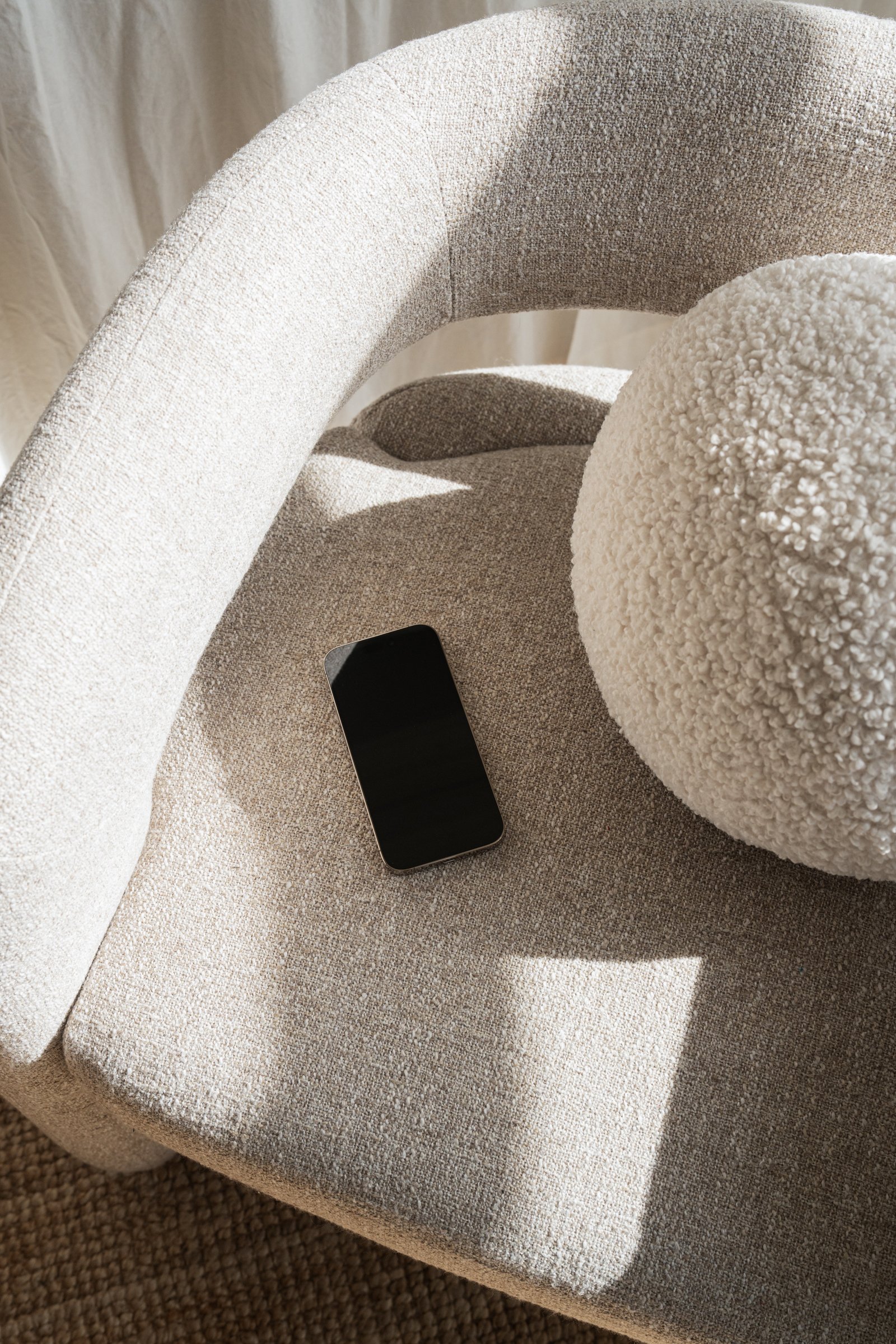 A beige upholstered armchair with a round, textured white pillow on it. A black smartphone is resting on the seat of the chair. Shadows and sunlight are visible on the chair and pillow.