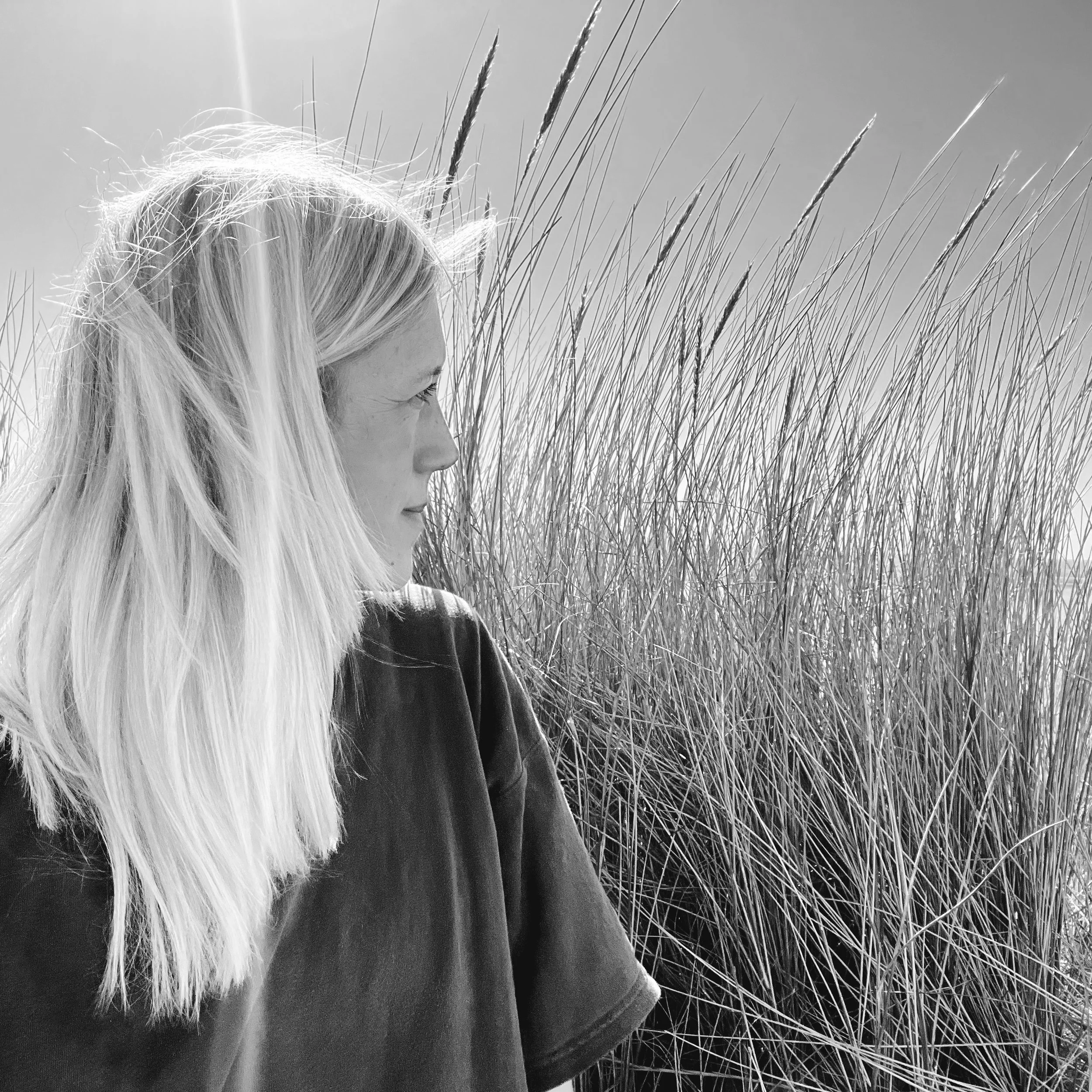 A woman with long blonde hair gazes to the left, standing in front of tall grass.