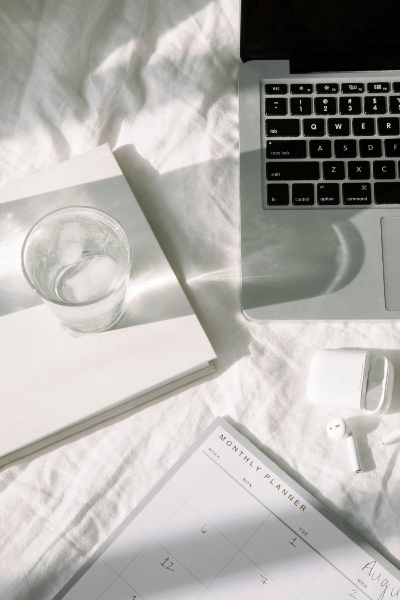 A workspace with a silver laptop, a transparent glass of water with ice, a white planner, a pair of wireless earbuds, and an Apple charger on a white textured surface with sunlight casting shadows.