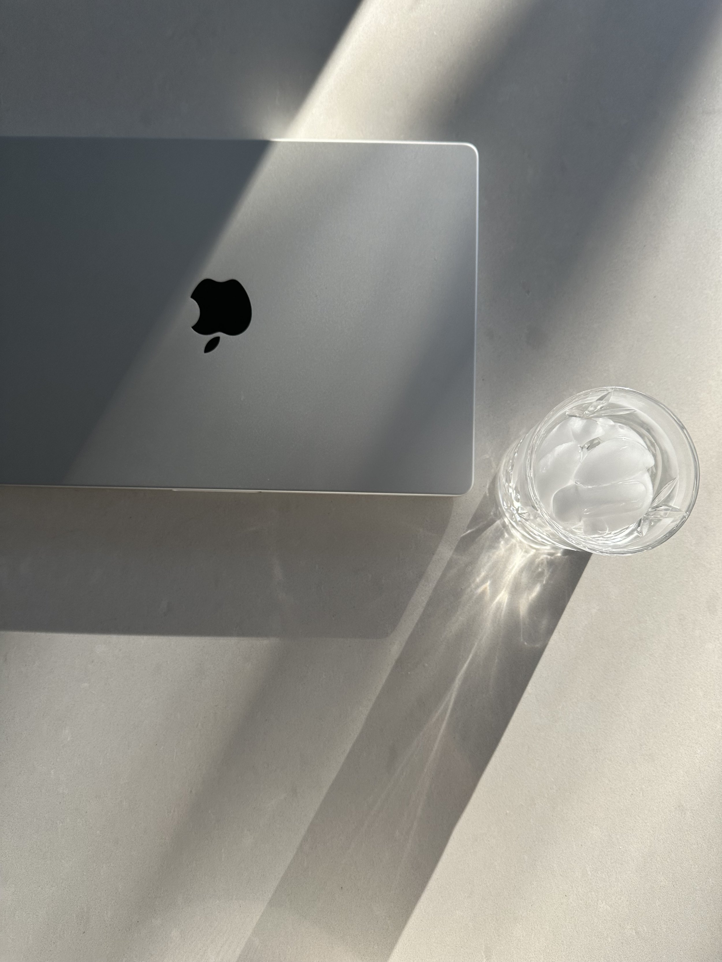 Top view of a closed Apple MacBook laptop on a light-colored surface with modern glass mug filled with ice cubes, casting shadows in natural light.
