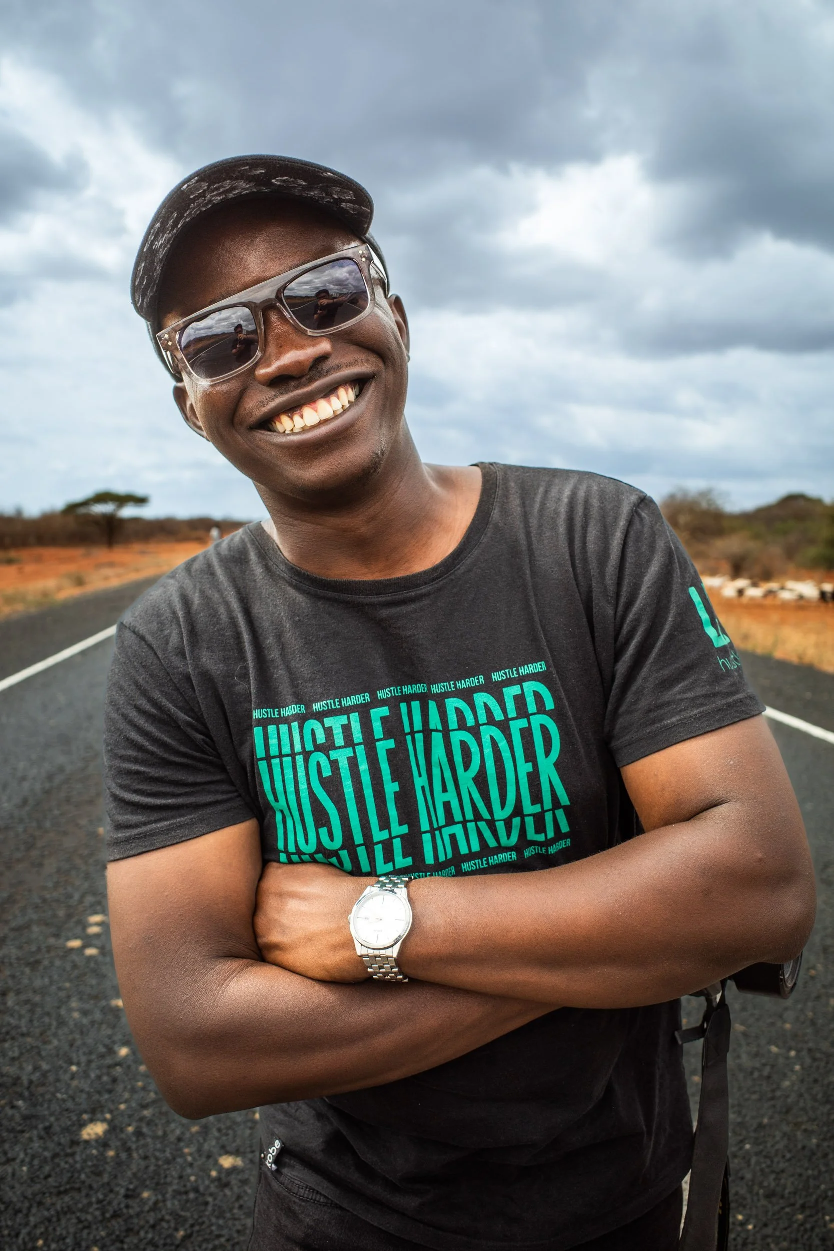 Paul Kidero wearing sunglasses, a black t-shirt with the words HUSTLE HARDER, a white wristwatch, and a black cap, standing on a rural road with a cloudy sky in the background.