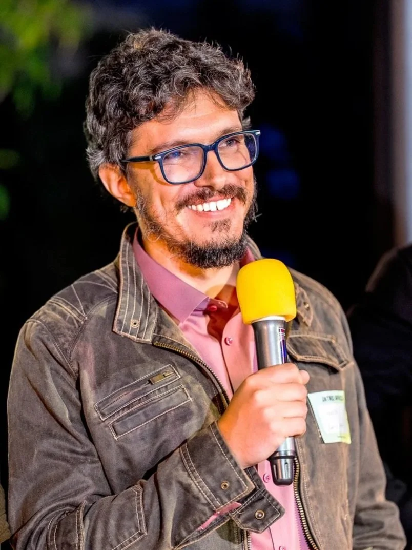 Man with curly hair, glasses, and a beard speaking into a yellow microphone, wearing a pink shirt and a brown jacket, smiling.