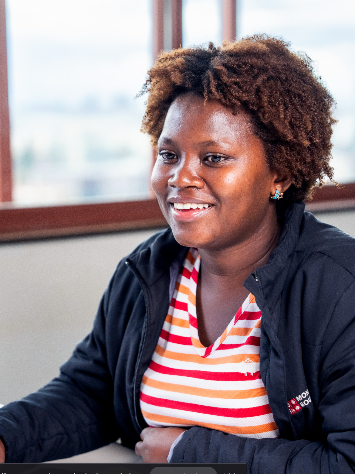 A woman with curly brown hair smiling, wearing a black jacket and a striped orange, red, and white shirt, sitting indoors near a window.