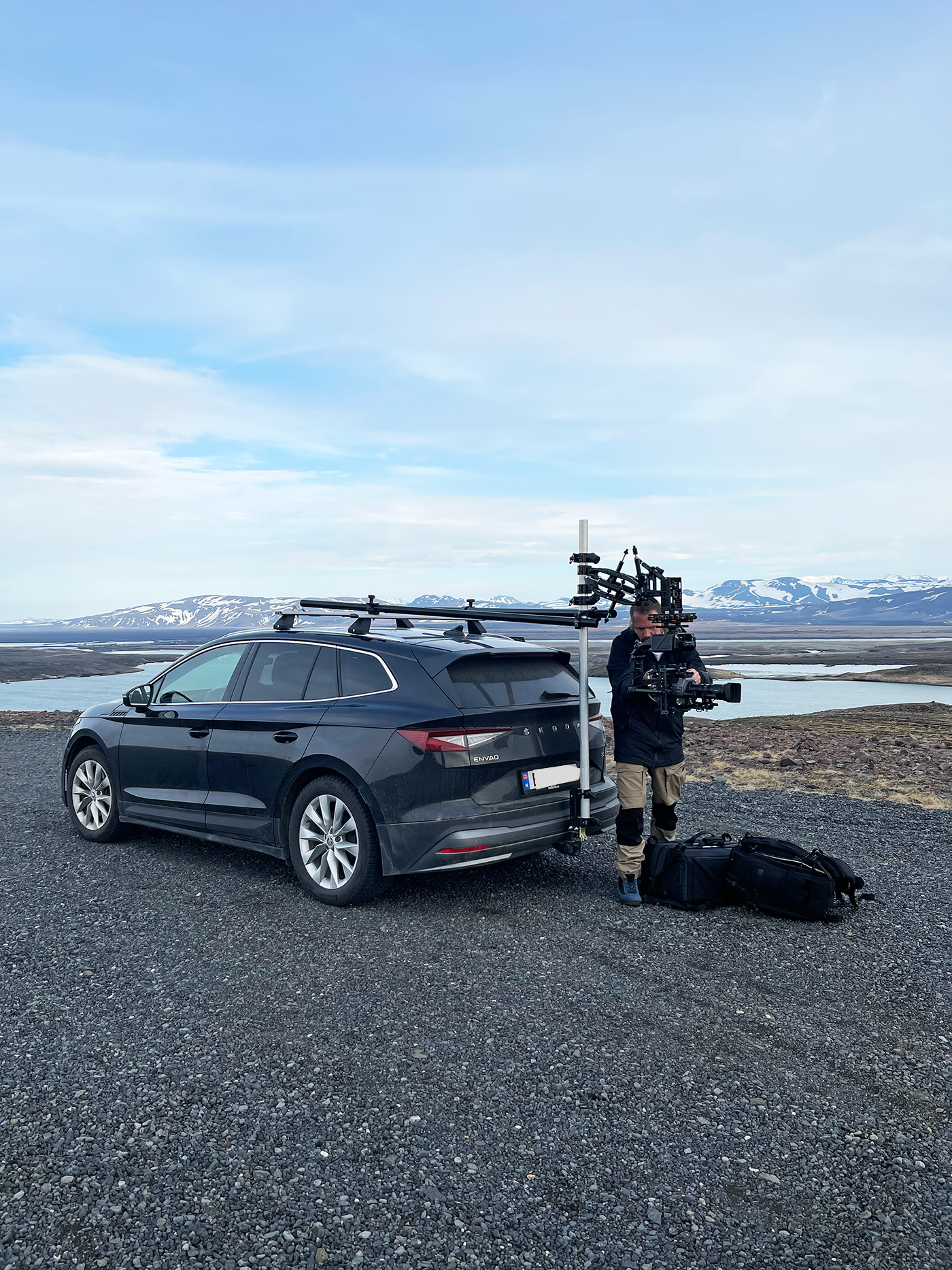Filmmaker with camera rig next to a dark gray station wagon on gravel, with mountains and a body of water in the background, under a partly cloudy sky.