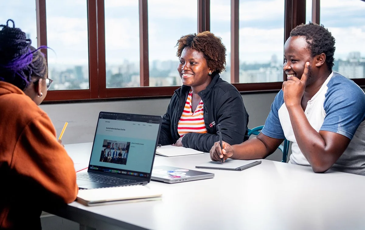 Three Thought Leader production members having a meeting at a table in a high-rise office with large windows and city views, smiling and engaged. Thougth Leader Nairobi Office.