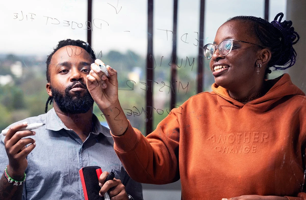 man and a woman, engaged in a discussion or brainstorming session. They are standing in front of a glass board with writing on it. The man holds a marker and looks directly at the camera, while the woman, wearing glasses and a brown hoodie, is writing on the glass board and smiling.