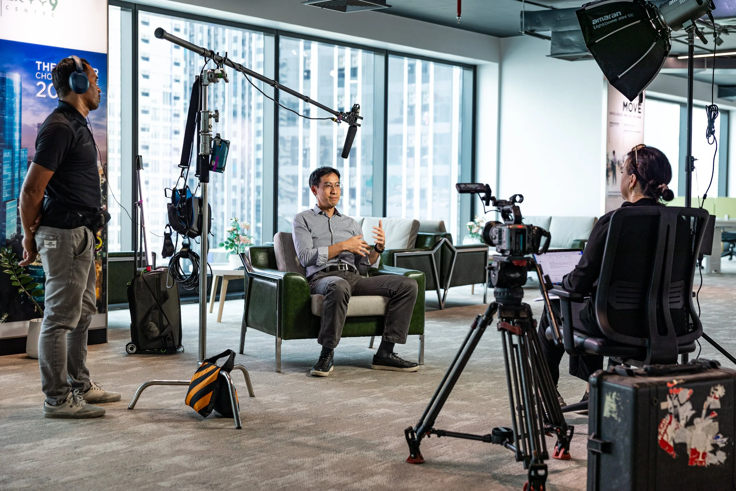 An interview or video recording session taking place in an office. A man in a gray striped shirt is seated on a green armchair, speaking and gesturing with his hands. A woman with an open laptop and headphones is filming him using a professional camera. A crew member with headphones is standing nearby, adjusting equipment. The setting is modern with large windows, cityscape views, and professional lighting.