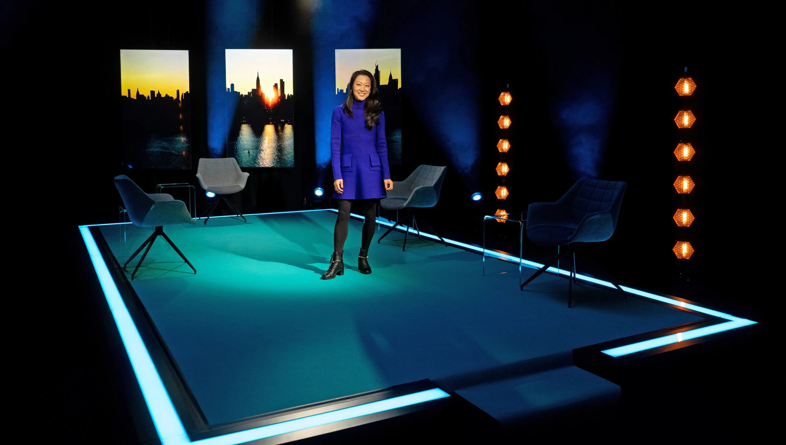 SHE Conference Host in a blue dress standing on the show set with colorful background images and four chairs around her.