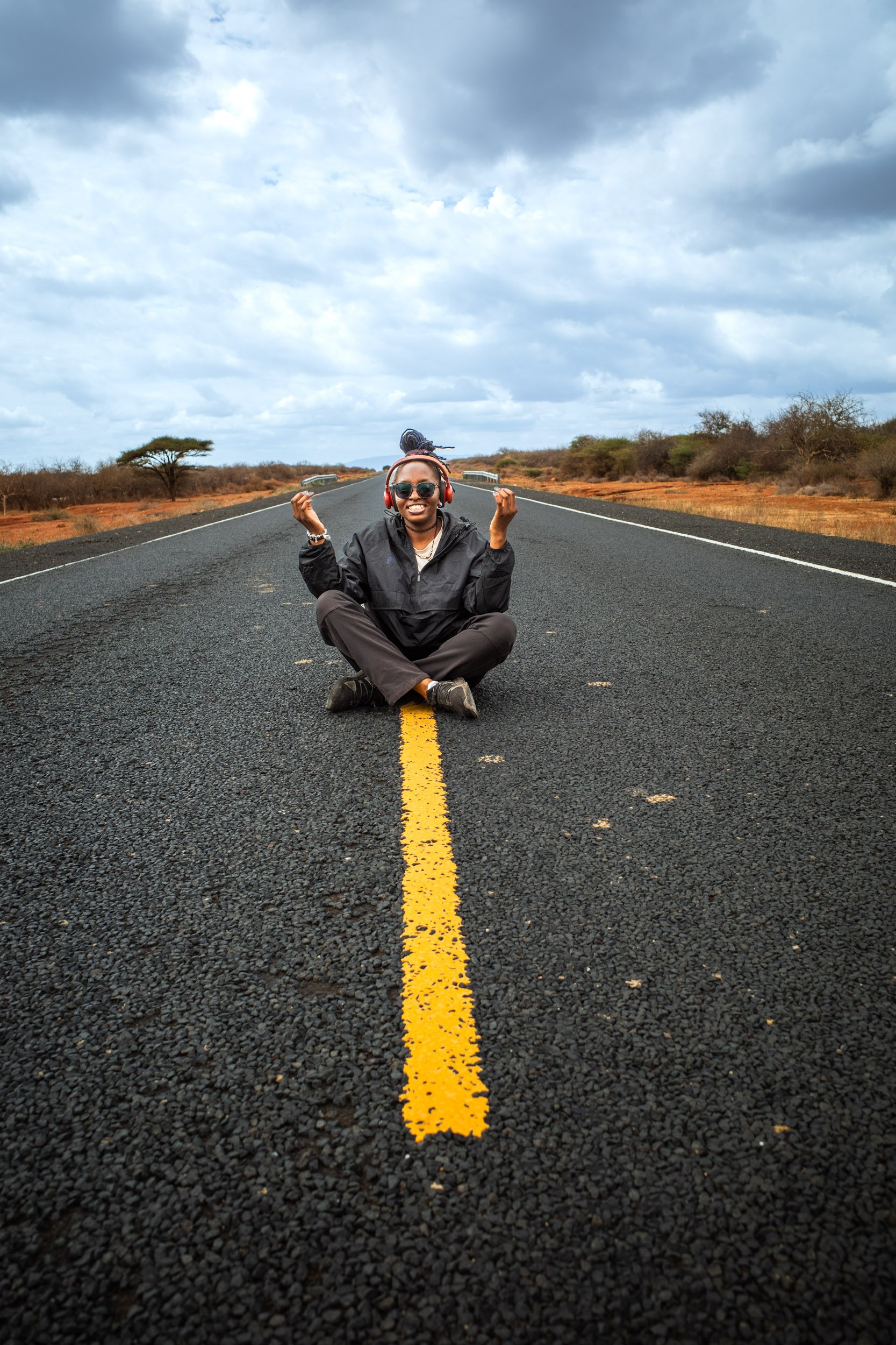 A woman sitting cross-legged in the middle of a remote road, smiling and wearing headphones, sunglasses, a black jacket, and dark pants, with a cloudy sky above and desert landscape on either side.