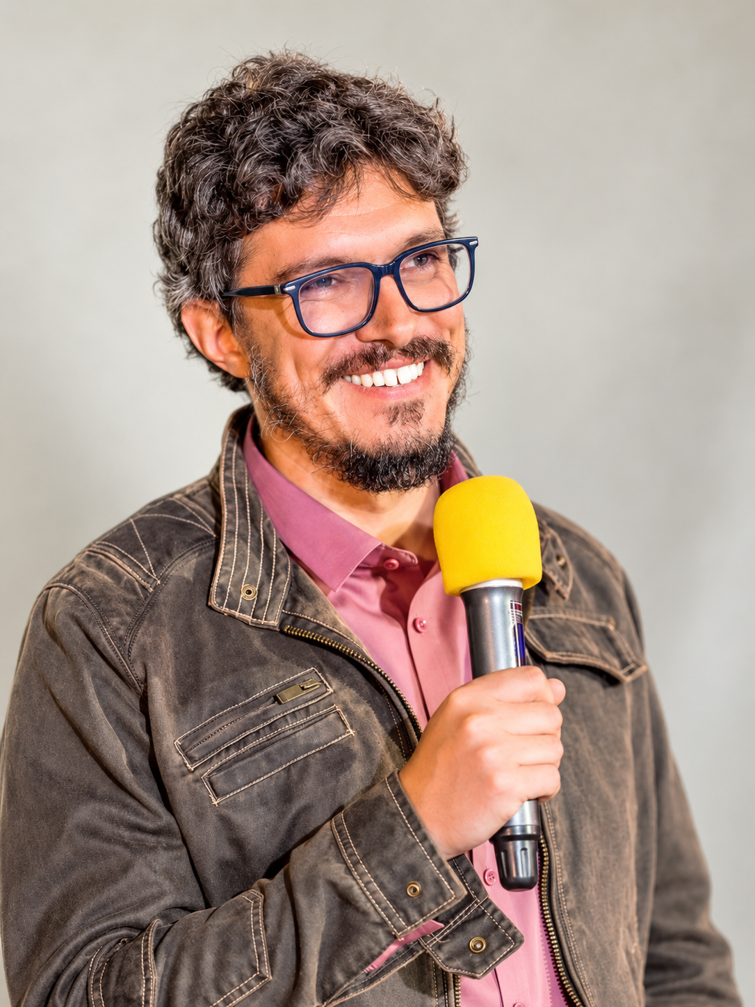 Man with curly hair, glasses, and a beard speaking into a yellow microphone, wearing a pink shirt and a brown jacket, smiling.