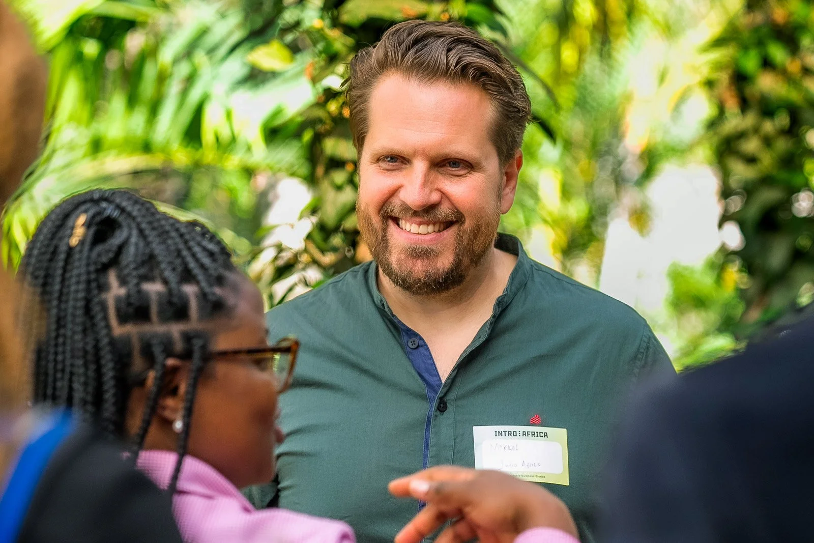 Mikkel Becker-Aakervik talking with a woman with braided hair and glasses at an outdoor event surrounded by green foliage.
