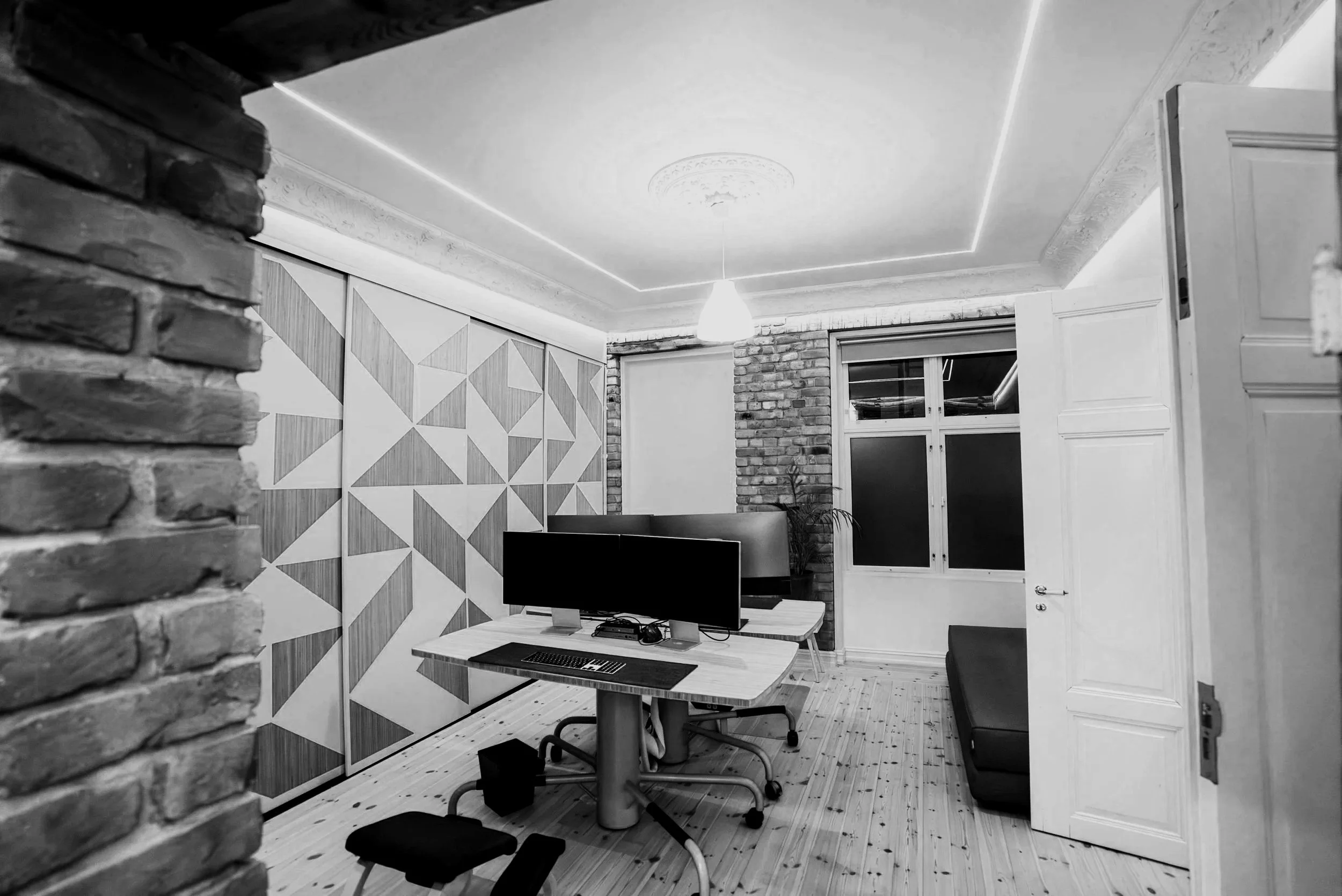Thought Leader office in Oslo, modern office with a wooden desk, dual monitors, a keyboard, and a chair, set against a geometrically patterned wall, featuring a sofa, brick wall, and window with blinds.