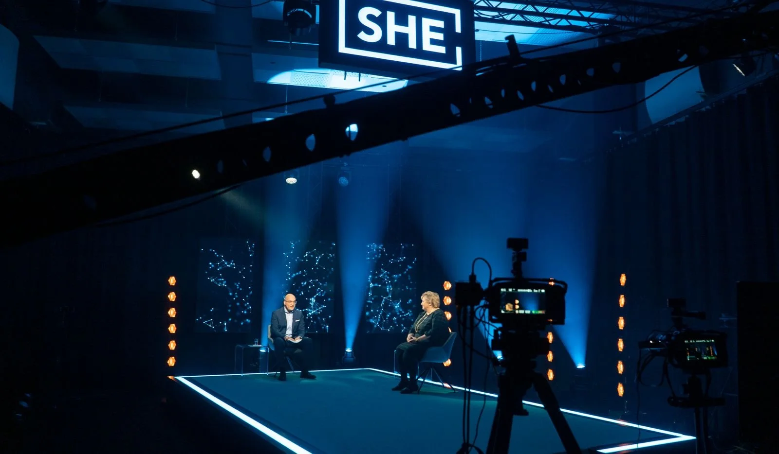 Two people are seated on the SHE stage during a recorded interview or discussion, with colorful digital graphics displayed behind them and lighting creating a professional studio atmosphere.